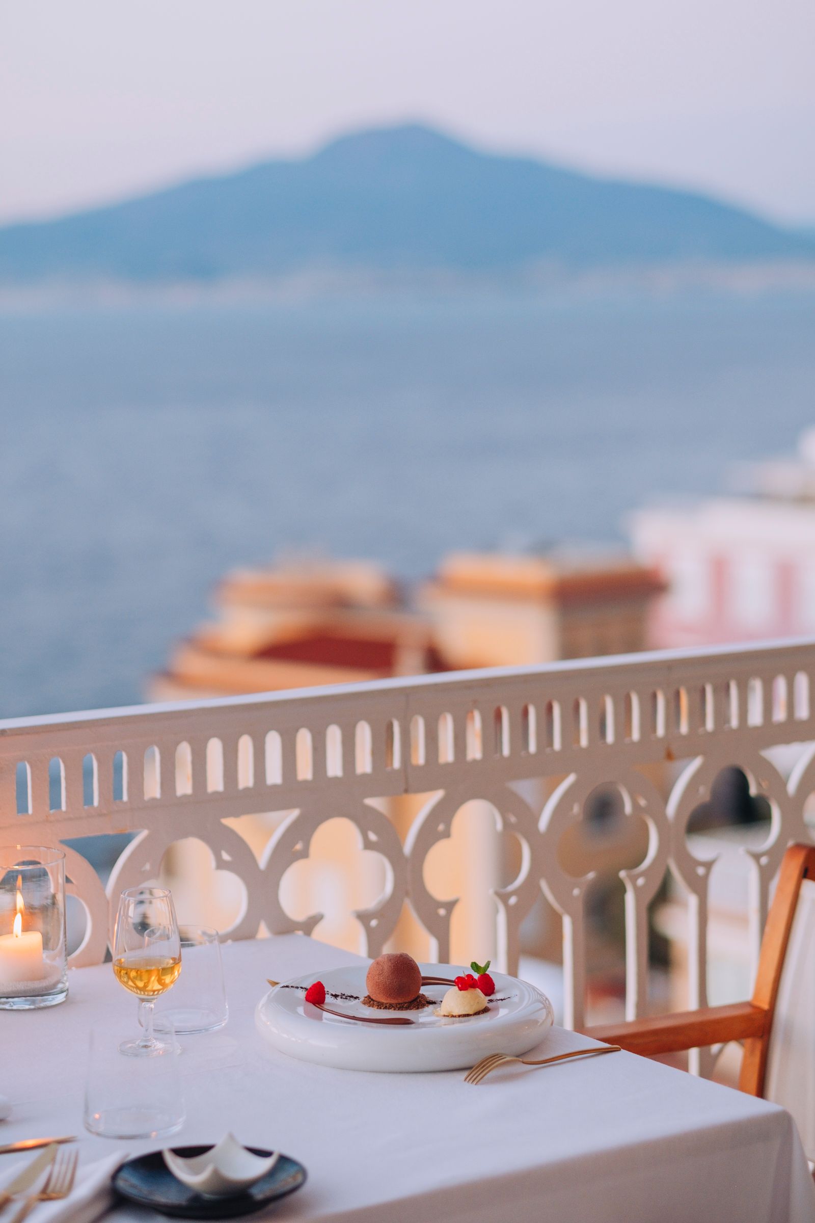 Un tavolo con dessert gourmet e vino, con vista sull'oceano e sul Vesuvio da un balcone al crepuscolo.