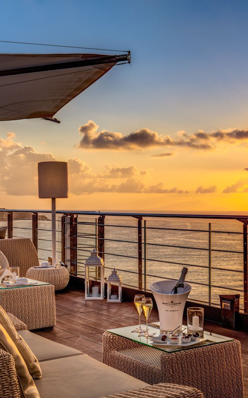 A sunset view from a deck with wicker lounge chairs, a champagne bucket, and railings overlooking the ocean.