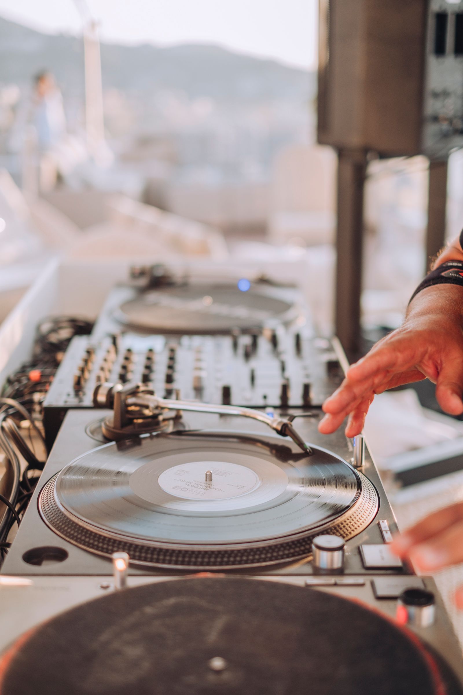 A DJ's hand adjusts a vinyl record on a turntable during an outdoor daytime event, with a mixer visible in the background.