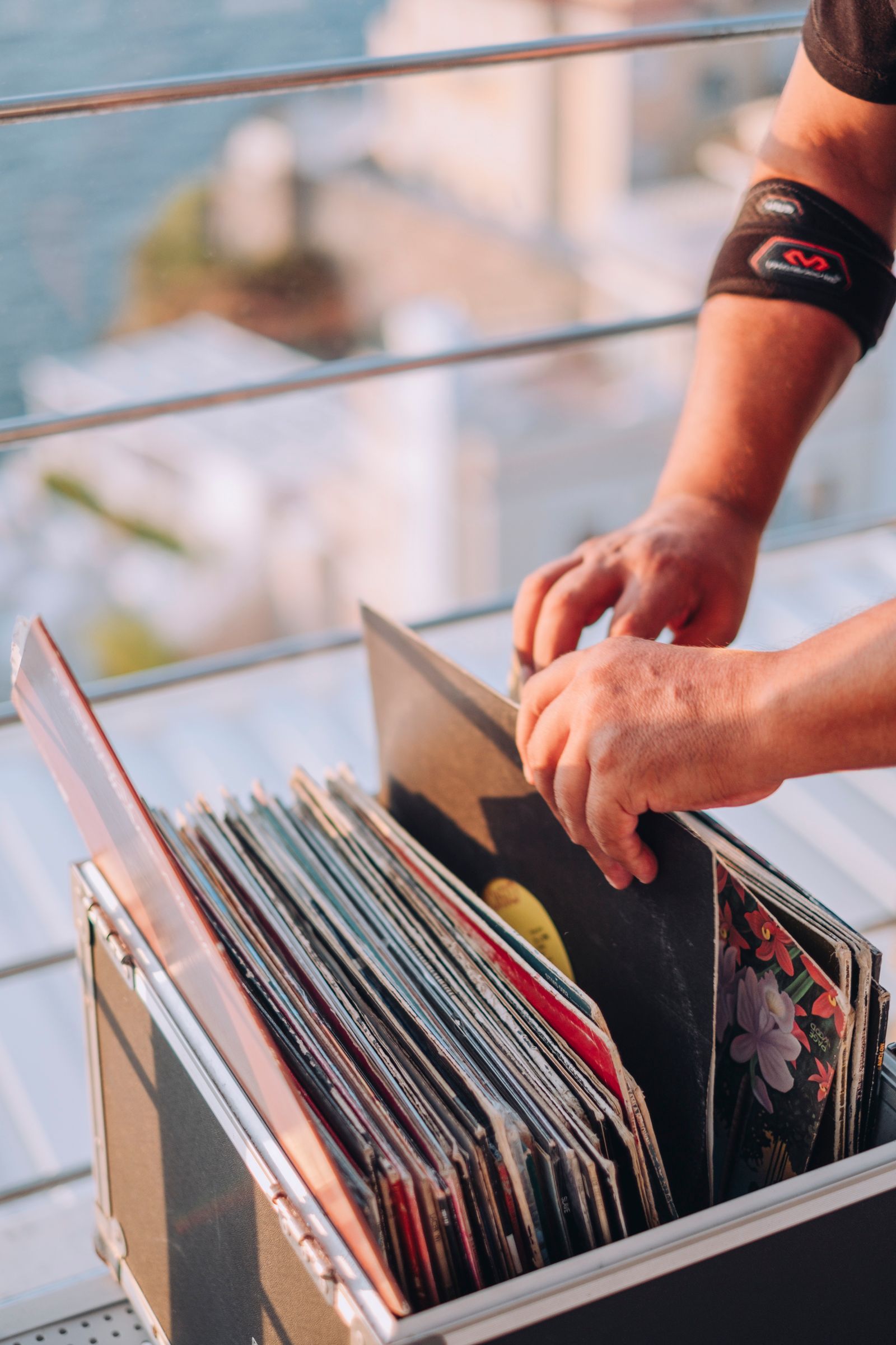 A person with an arm brace selecting a vinyl record from a carrying case on a balcony overlooking the water.