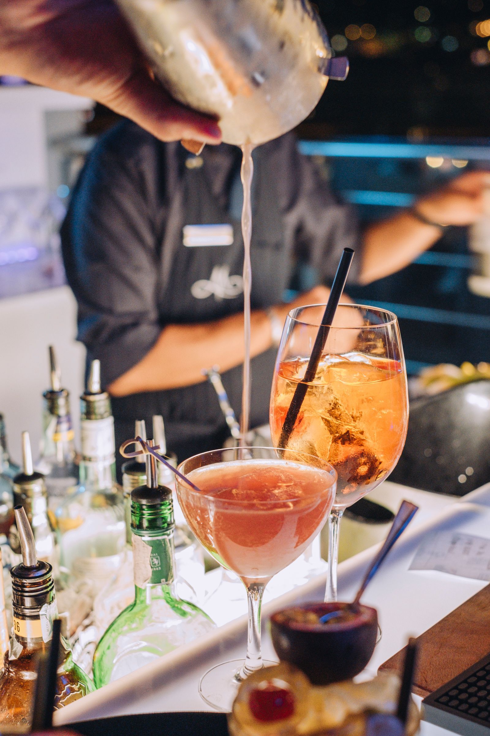 In un bar affollato e illuminato da luci soffuse, un barista versa un cocktail da uno shaker di metallo in un bicchiere.