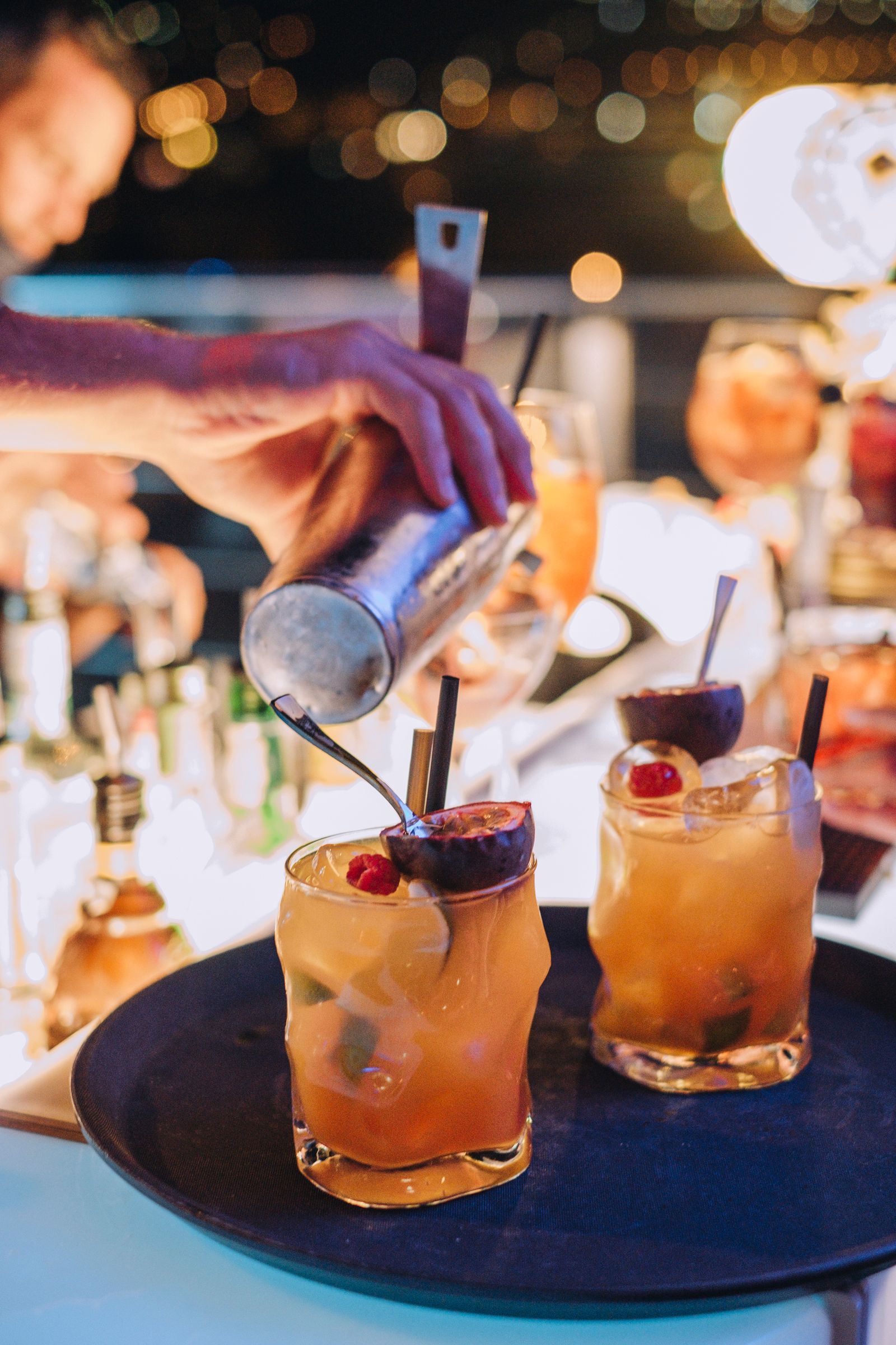 Un barista versa un cocktail in un bicchiere posto su un vassoio insieme a un'altra bevanda, sullo sfondo di un paesaggio serale all'aperto.