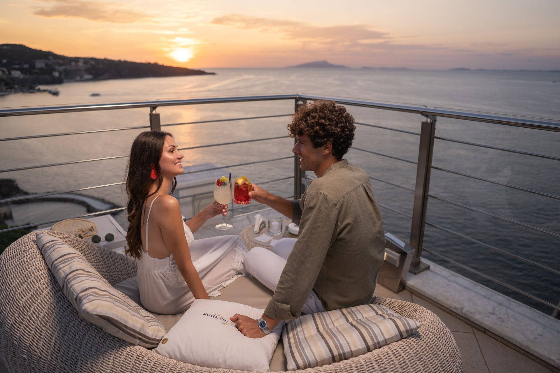 A couple toasts with cocktails on a sunset balcony overlooking the ocean.