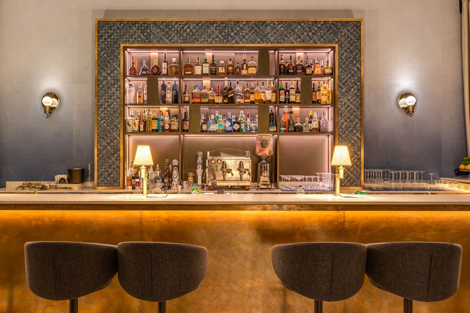 Un elegante bancone bar con una base metallica luminosa, quattro sedie grigie e uno scaffale retroilluminato pieno di bottiglie di liquori di vario tipo.