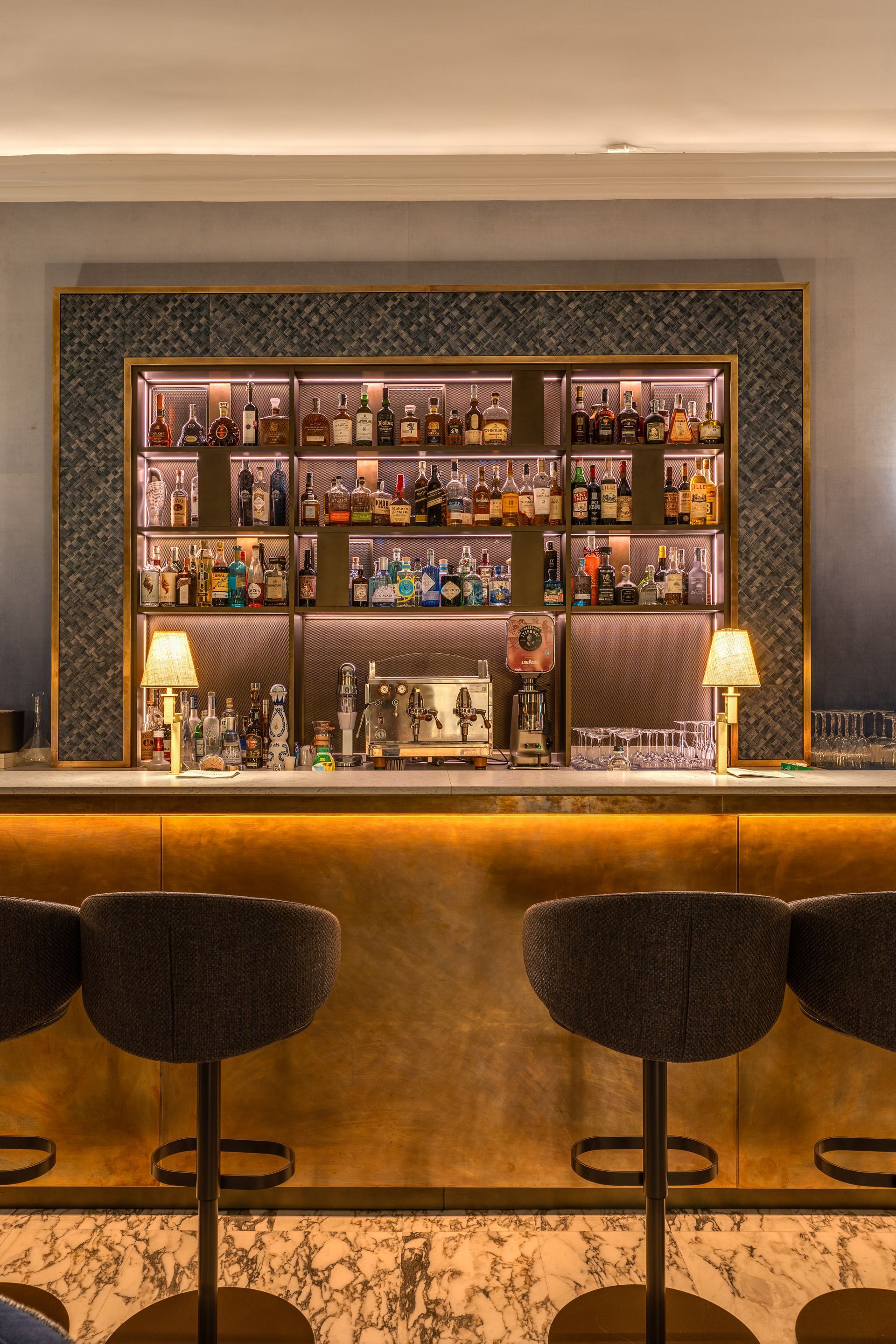 Un elegante bar d'hotel con sgabelli, una mensola illuminata con bottiglie in mostra e una calda illuminazione sotto il bancone, il tutto in un ambiente moderno.