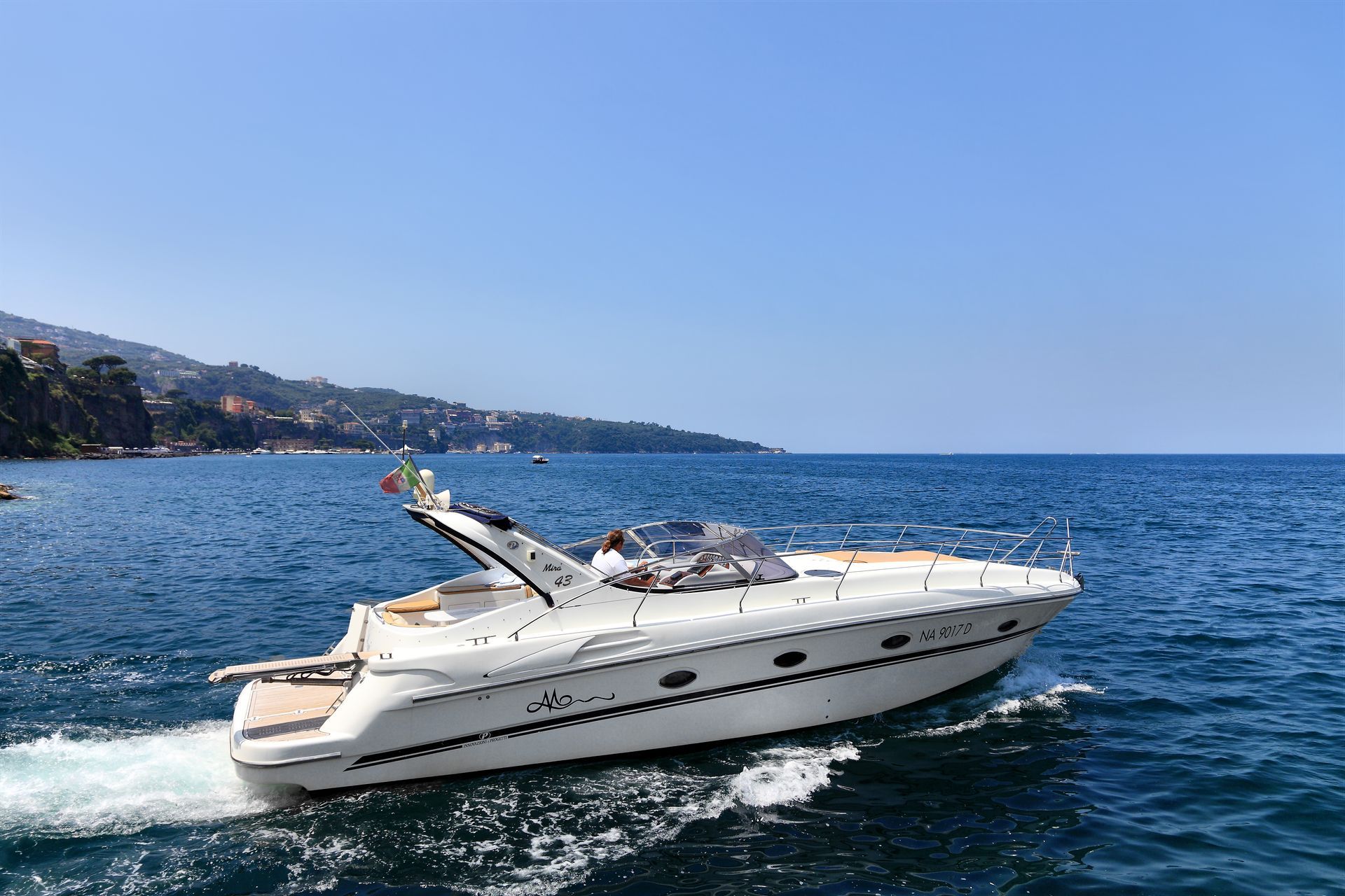 Un motoscafo bianco che naviga su acque blu calme, con una costa pittoresca sullo sfondo, sotto un cielo limpido.