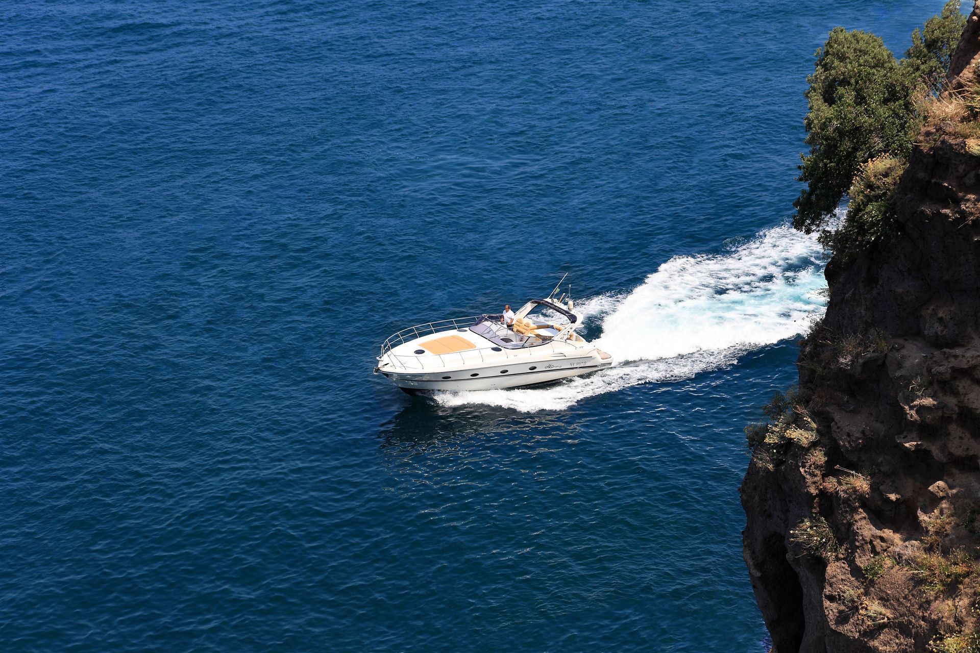 A white motorboat speeds across deep blue water near a rocky cliffside, leaving a white wake behind it.