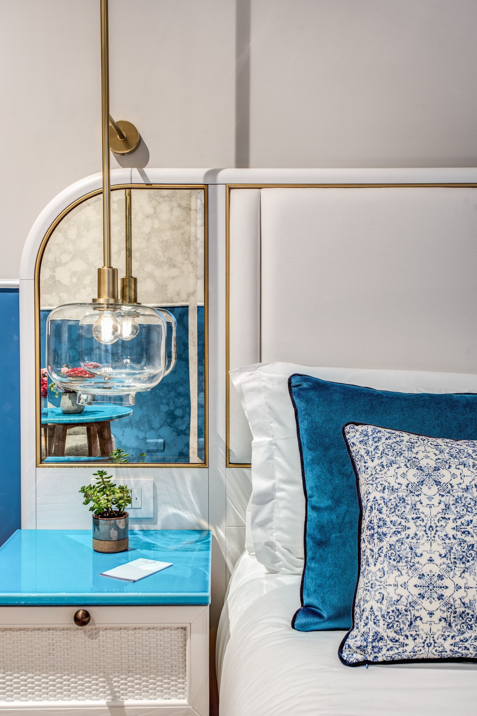 A white bedside table with a teal top, a small plant, and a glass pendant light by a bed with blue and patterned pillows.