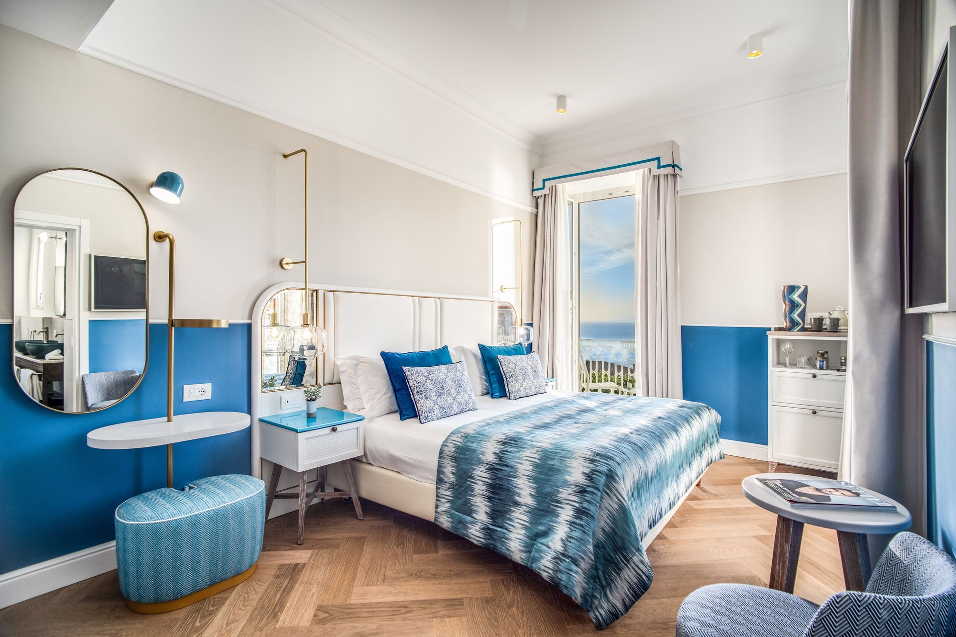 Una luminosa camera d'albergo con letto matrimoniale, lenzuola blu a fantasia, pavimento in legno, specchio a parete e balcone con vista mare.