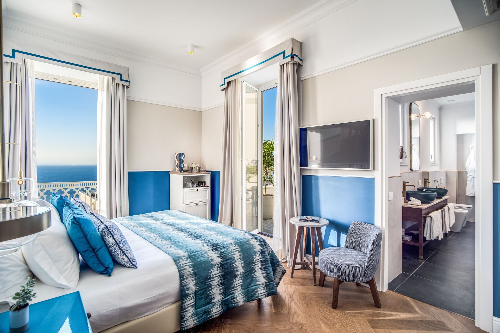 Una camera d'albergo con un letto a fantasia blu e bianca, una sedia, un tavolino e un bagno con vista sull'oceano.