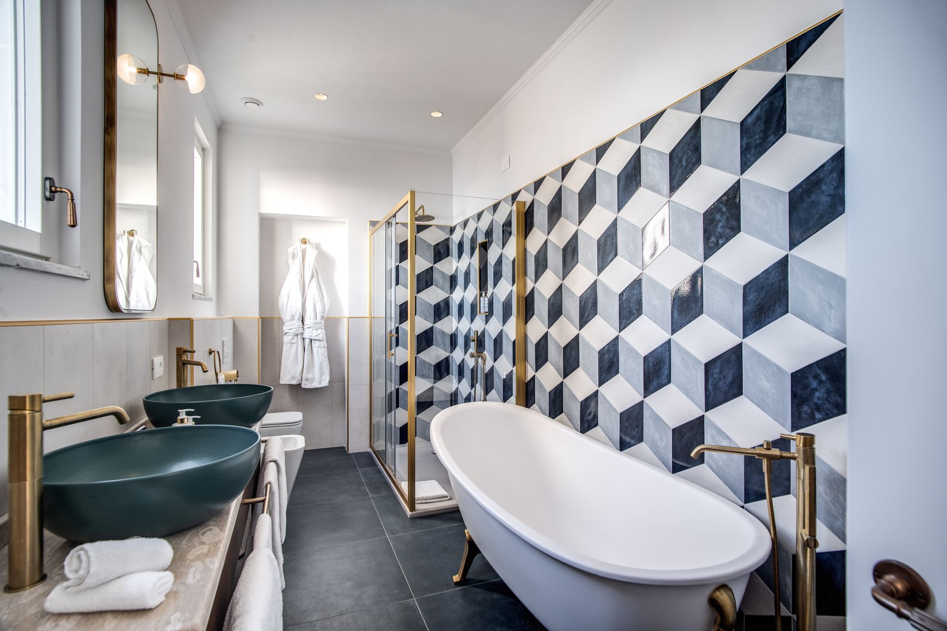 Un bagno con lavabi a ciotola verde scuro, rubinetteria dorata, una vasca da bagno bianca con piedini e una parete rivestita con piastrelle geometriche blu e bianche.