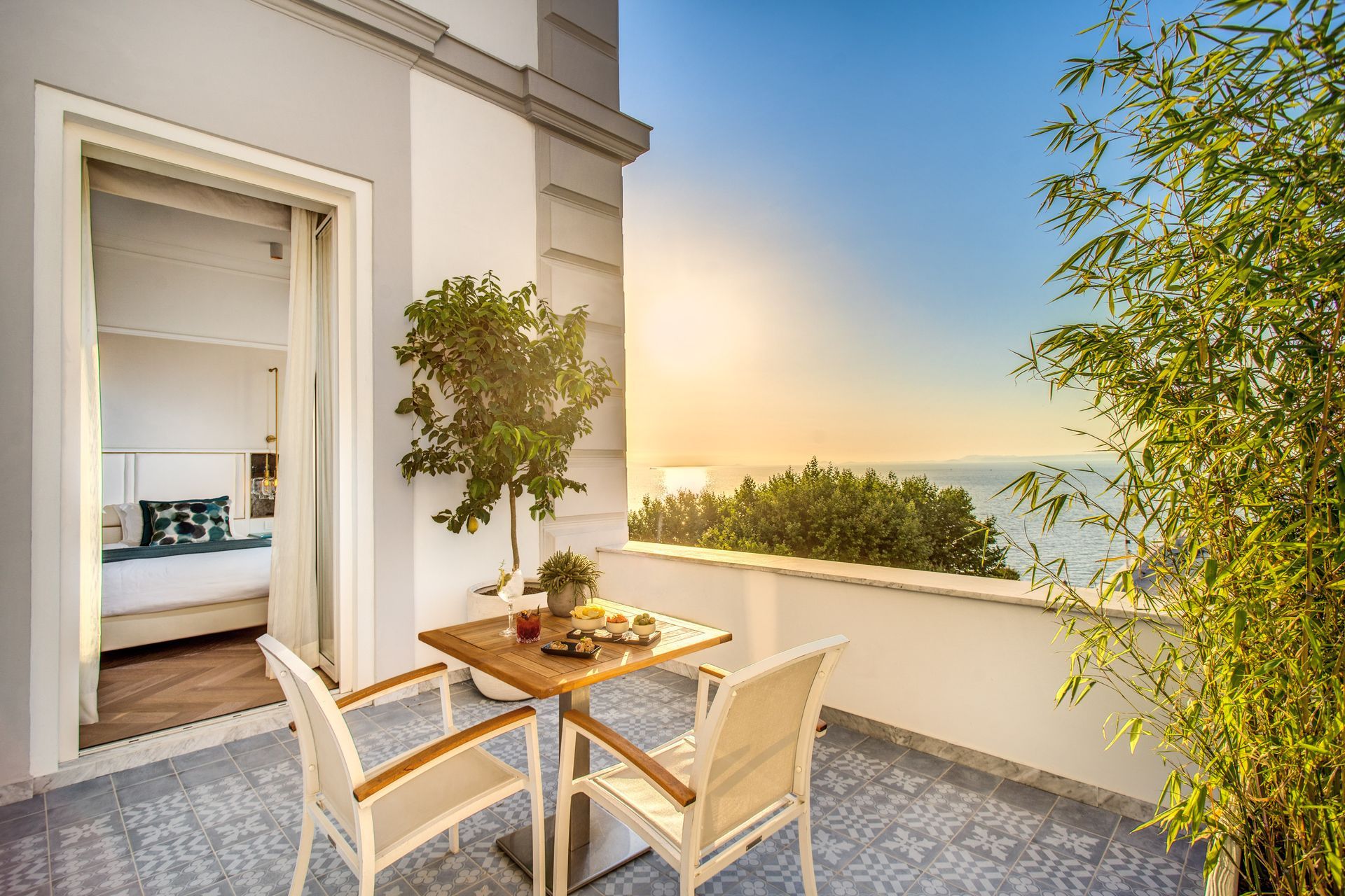 Un balcone al tramonto con un tavolino, due sedie e piante in vaso che si affaccia su un mare calmo.