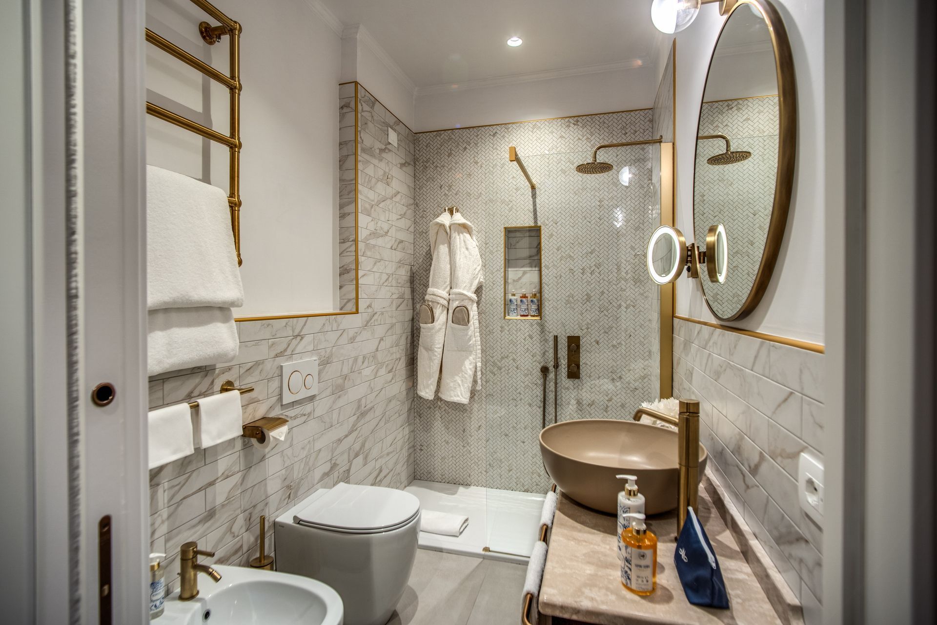 Un bagno moderno e lussuoso con pareti rivestite in marmo bianco, uno specchio rotondo, un rubinetto dorato, una cabina doccia e un accappatoio.