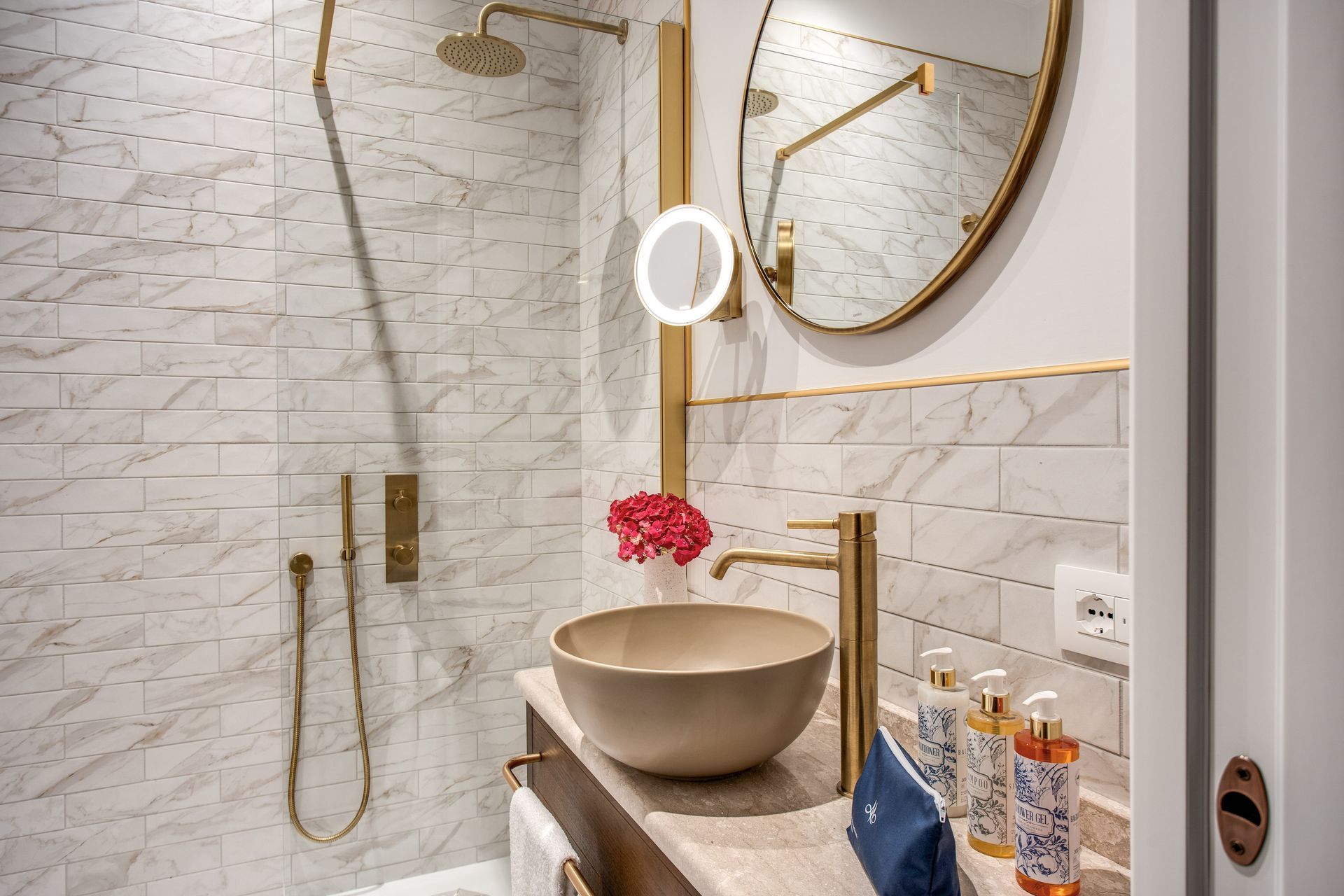 Un bagno moderno con lavabo a bacinella, rubinetto dorato, specchio rotondo e cabina doccia con piastrelle effetto marmo.