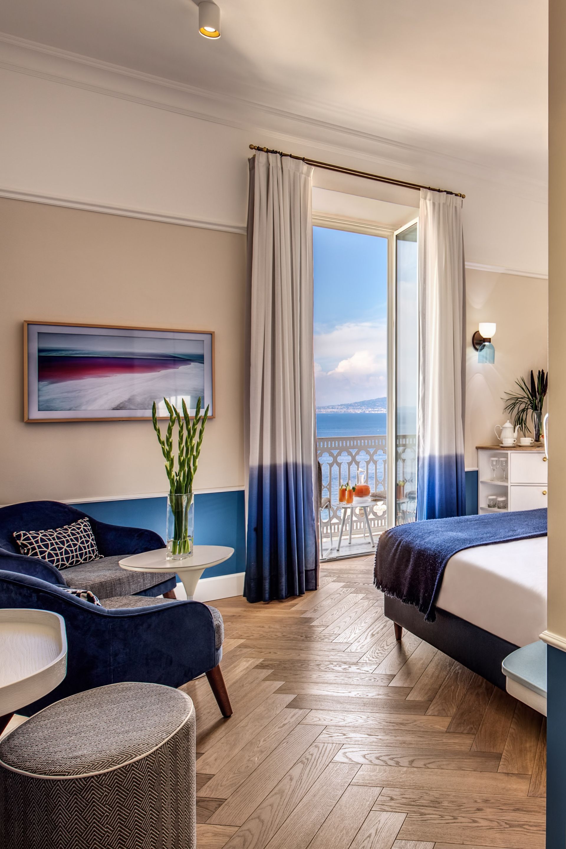 Una camera da letto con dettagli blu, pavimento in legno a spina di pesce e una porta-finestra che si affaccia sull'oceano.