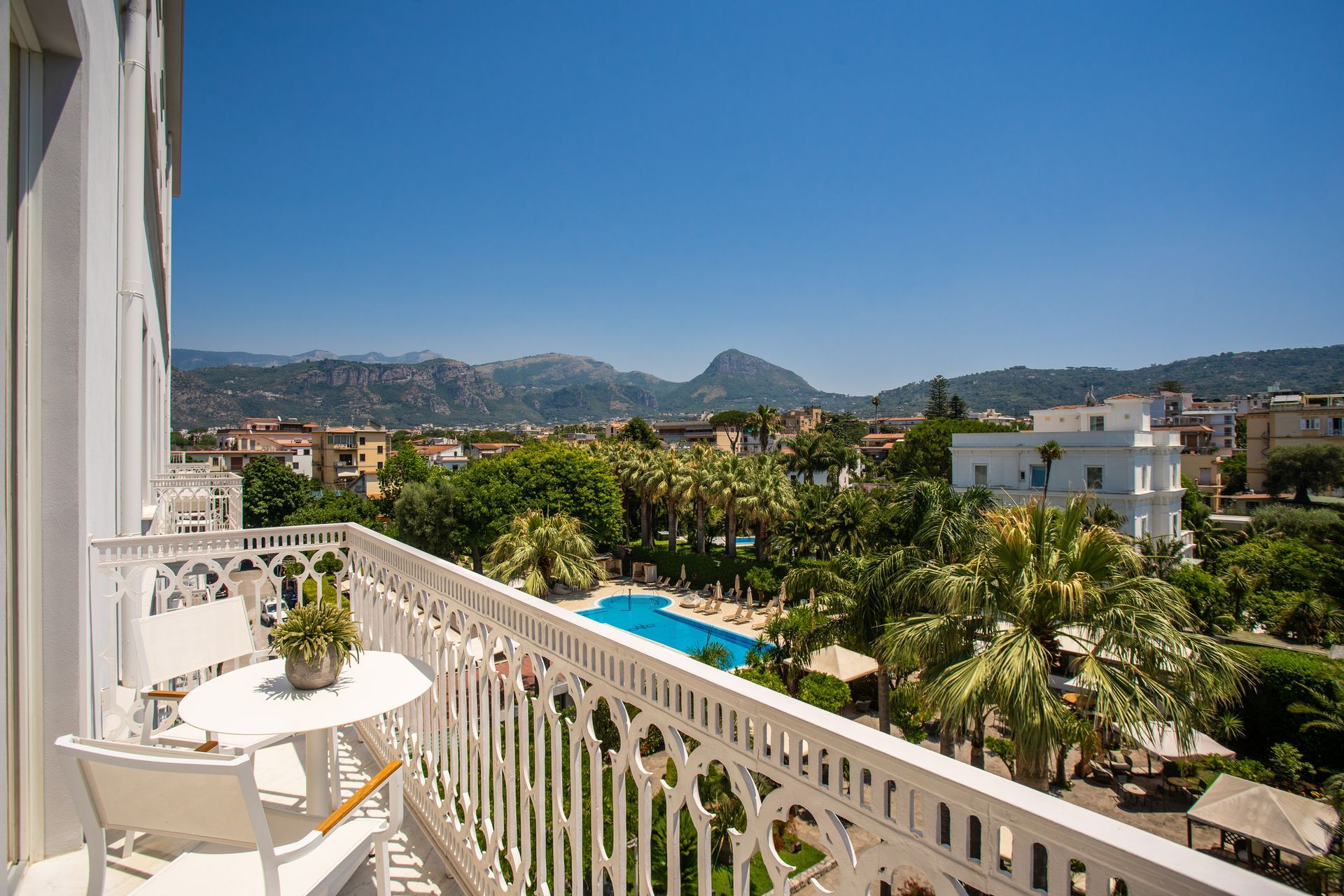 Dal balcone si gode di una vista sul giardino del resort, con piscina, palme e montagne in lontananza, sotto un cielo azzurro e limpido.