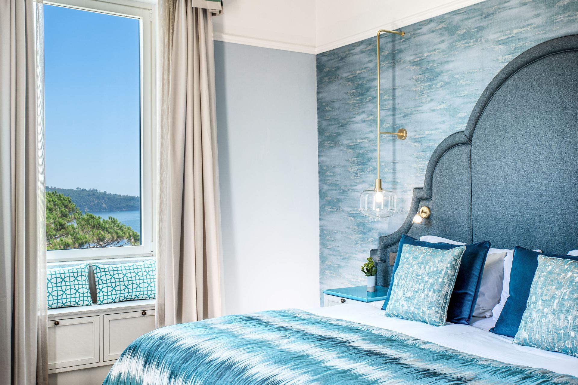Una lussuosa camera da letto con arredi blu, un'ampia testiera del letto e una finestra che si affaccia su uno splendido paesaggio costiero.