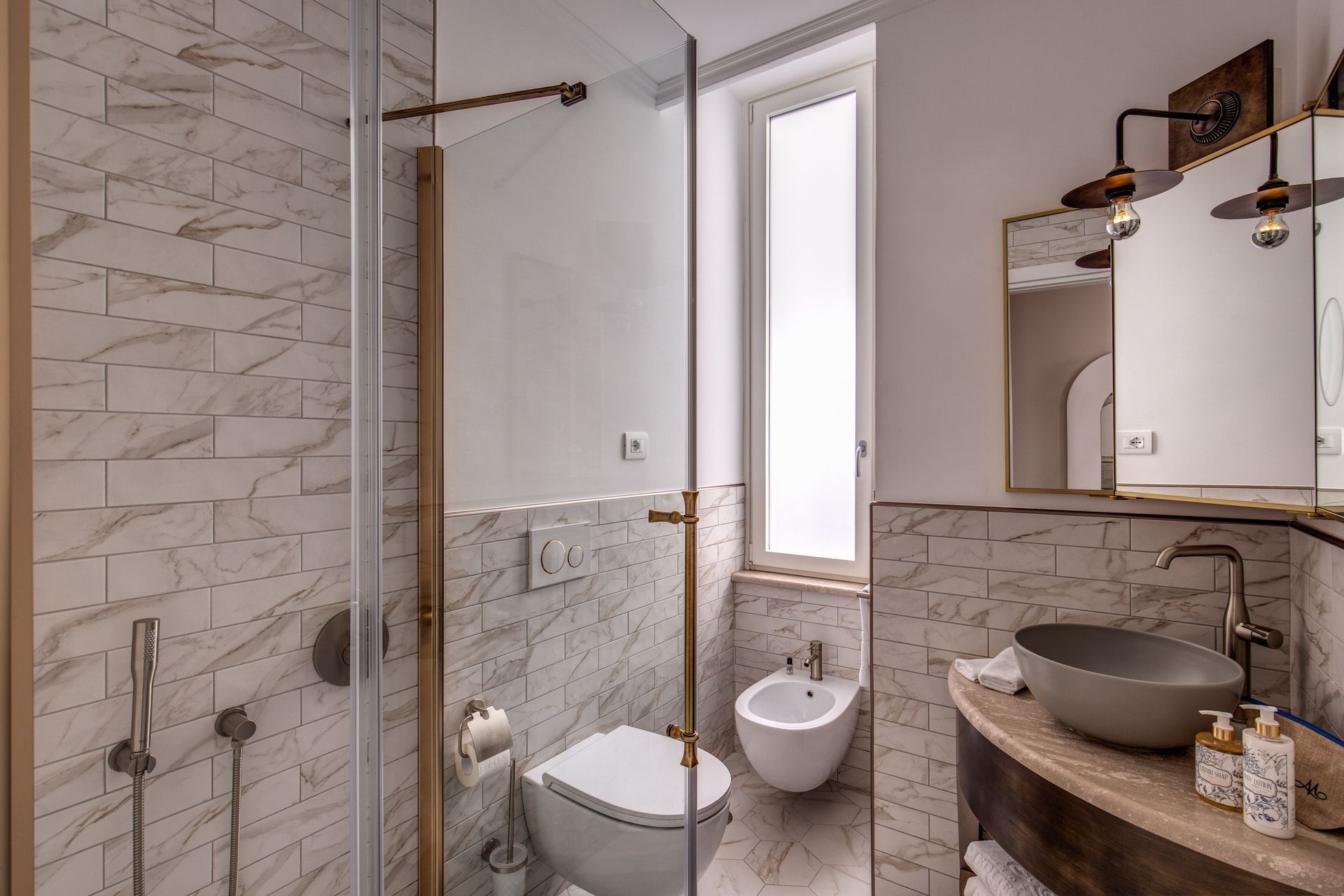 Un bagno moderno con pareti rivestite in marmo, cabina doccia, WC, bidet e lavabo a bacinella su mobiletto in legno.