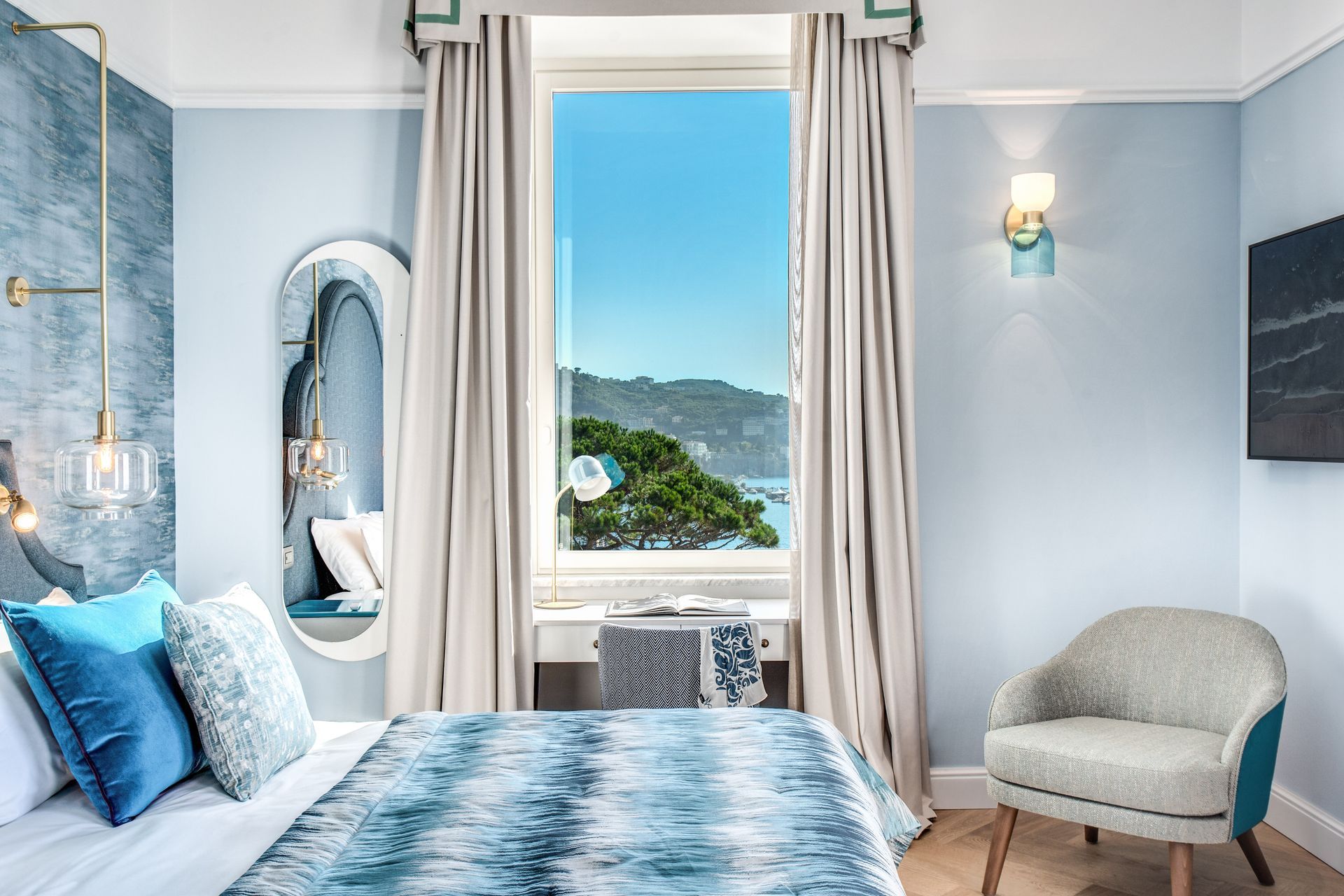 Una camera da letto azzurra con un letto con lenzuola a fantasia, uno specchio, una finestra con vista sulla costa e una poltrona beige.