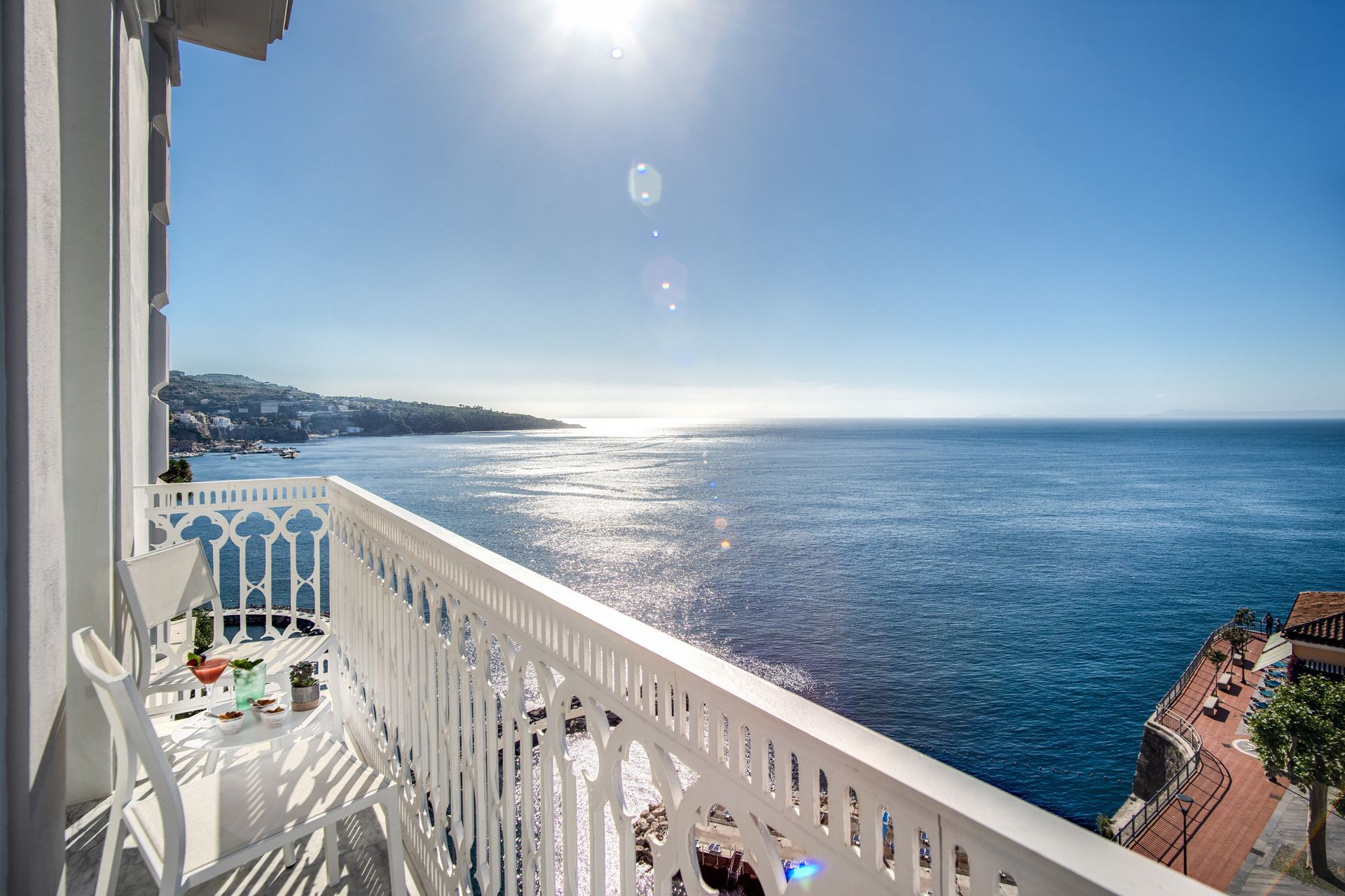 Un balcone bianco si affaccia su un calmo oceano blu sotto un cielo limpido e soleggiato, con una lontana cittadina costiera all'orizzonte.