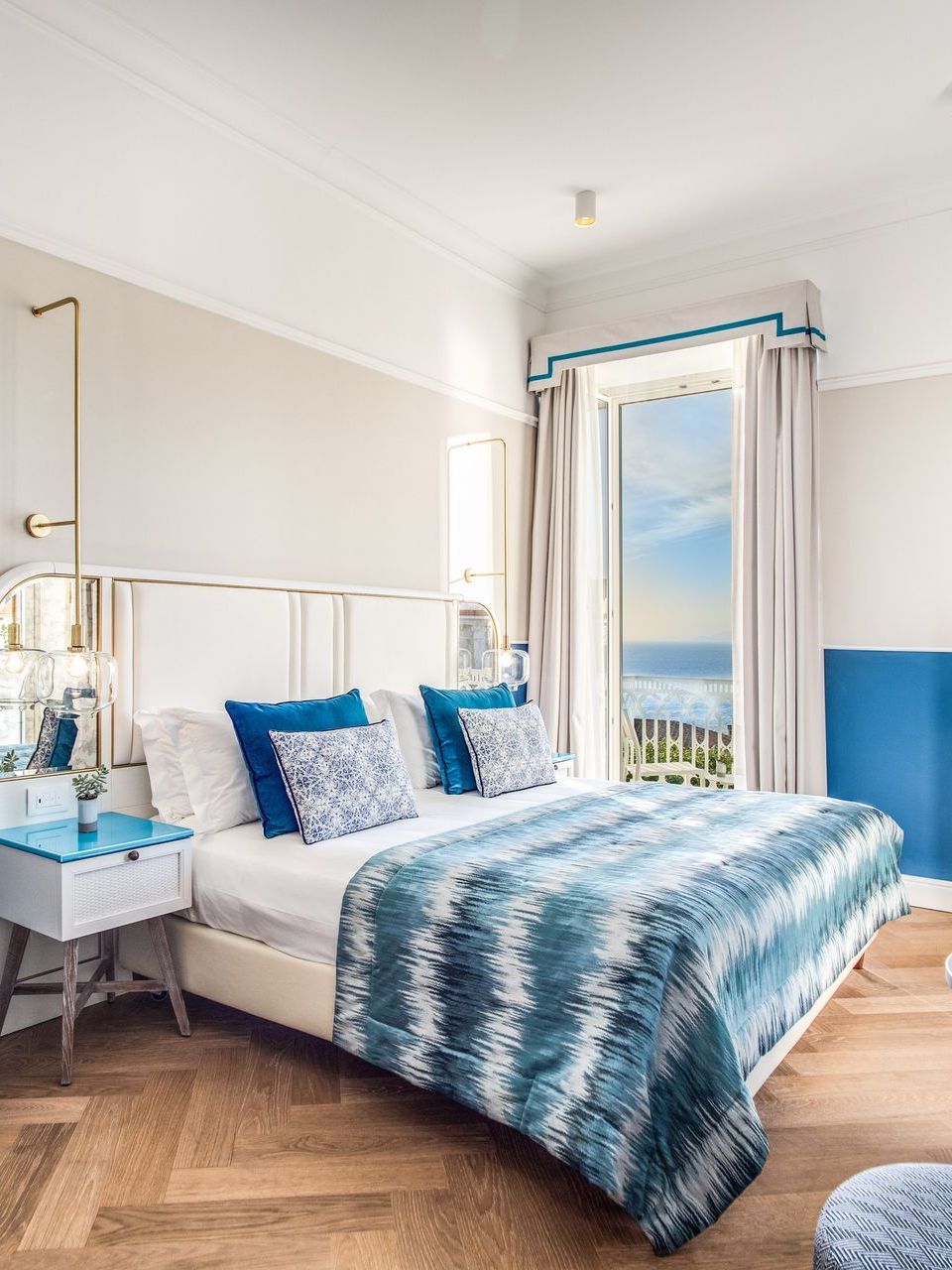 A bedroom with a queen bed featuring blue and white patterned bedding, a white nightstand, and a sea-view balcony.