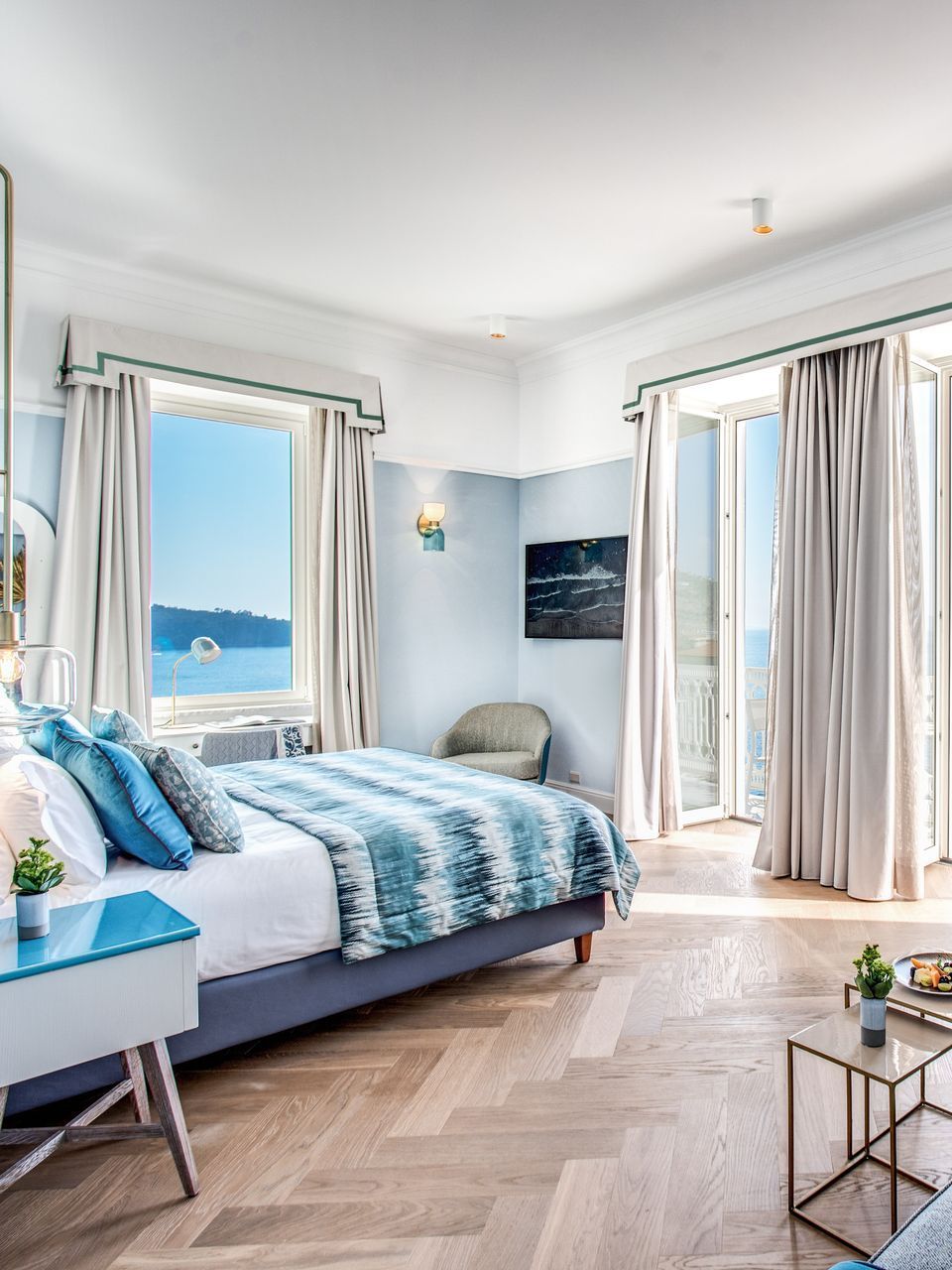 A bright, airy bedroom with a blue and white patterned bed, hardwood floors, and two large windows overlooking the sea.