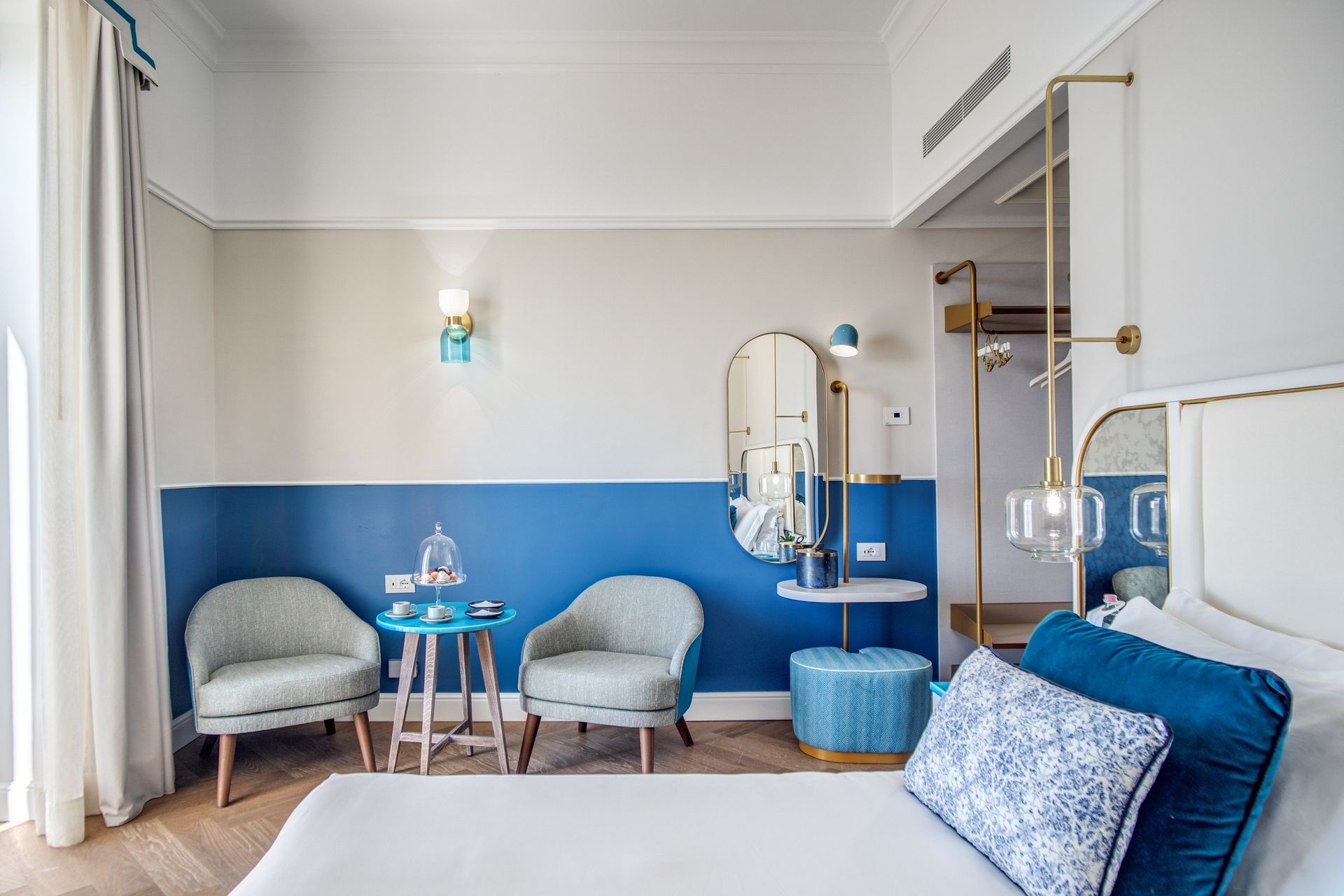 Una camera d'albergo luminosa e moderna, arredata con due poltrone grigie, un tavolino blu, uno specchio da trucco e un letto soffice.