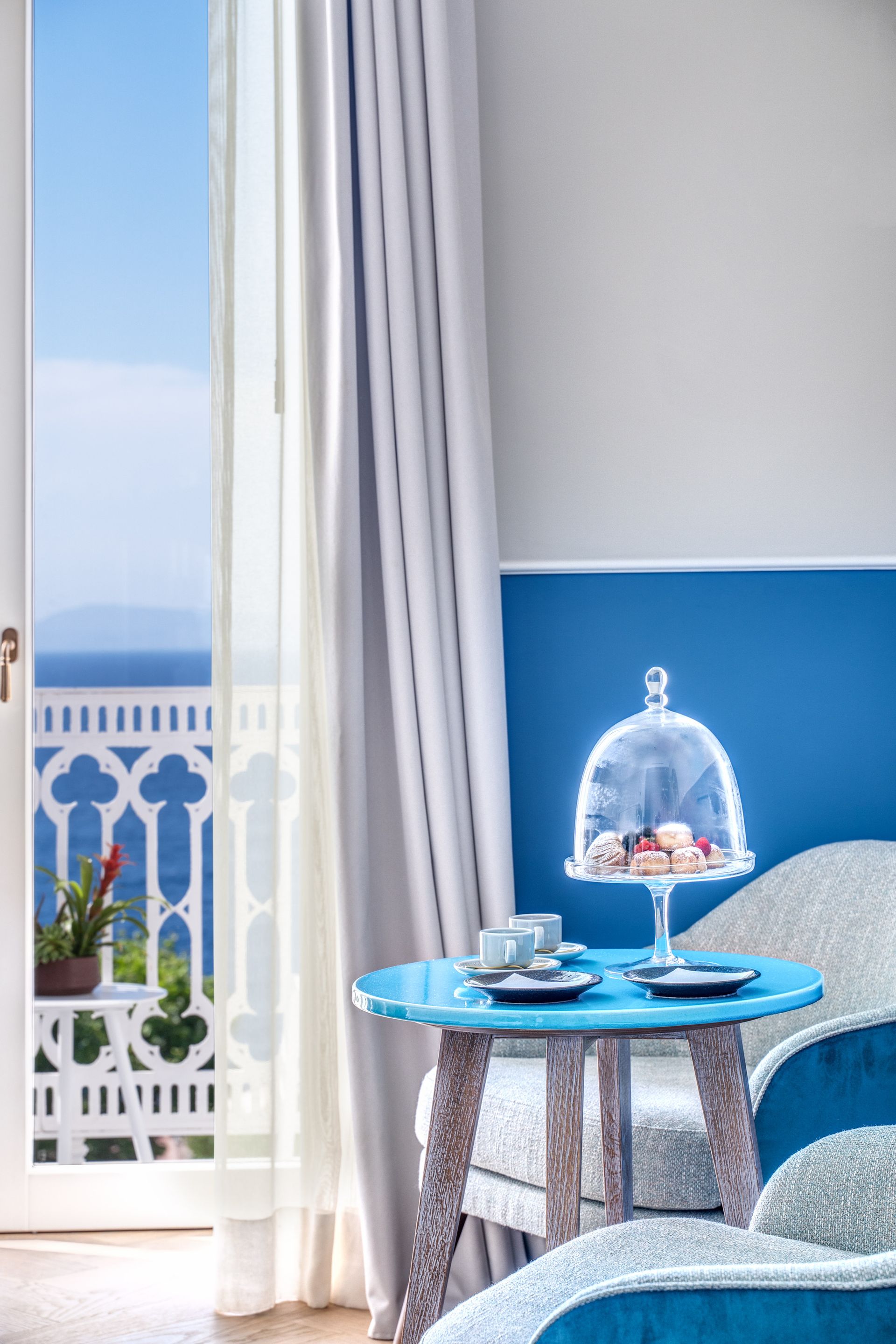Una stanza luminosa con pareti blu, un tavolino con tè e una campana di vetro, affacciata su un balcone con vista mare.