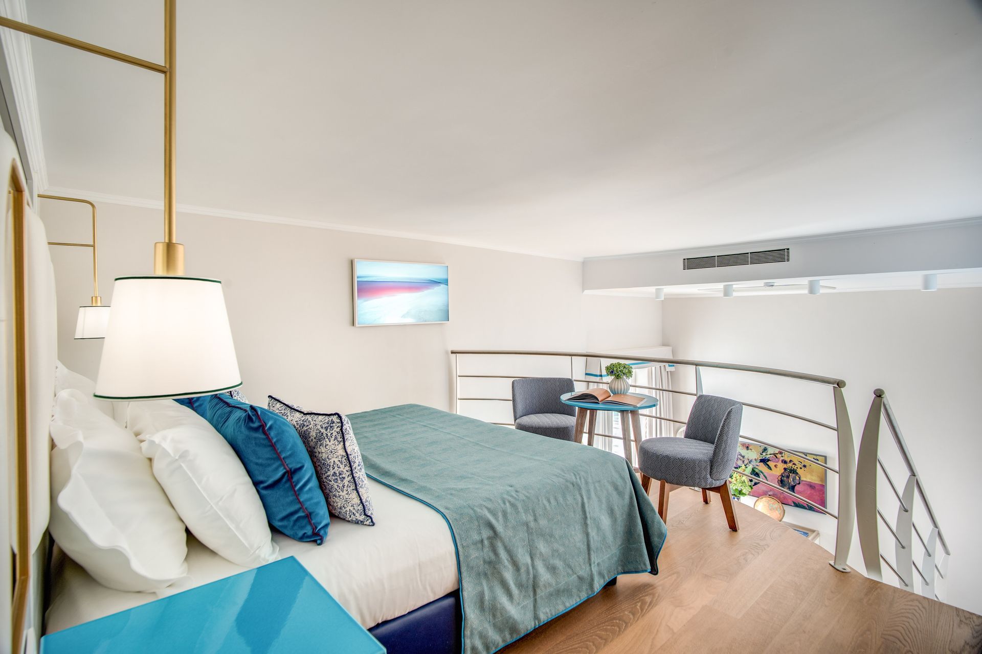 Una camera da letto in stile loft d'albergo con un letto bianco, una coperta color verde acqua, due sedie grigie, un tavolino e un televisore a parete.