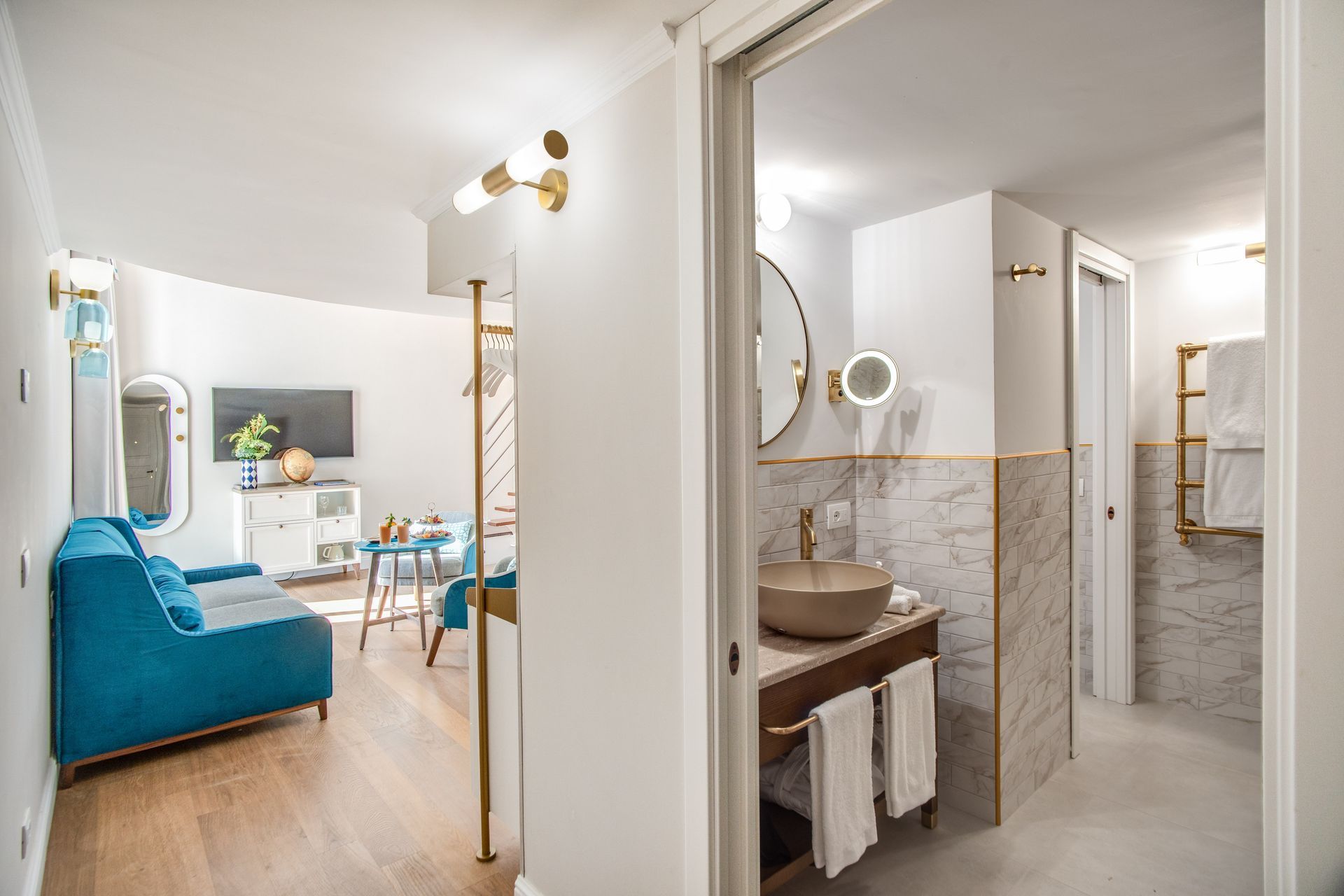 Una luminosa camera d'albergo con un divano blu e un bagno con lavabo in marmo e rubinetteria dorata.