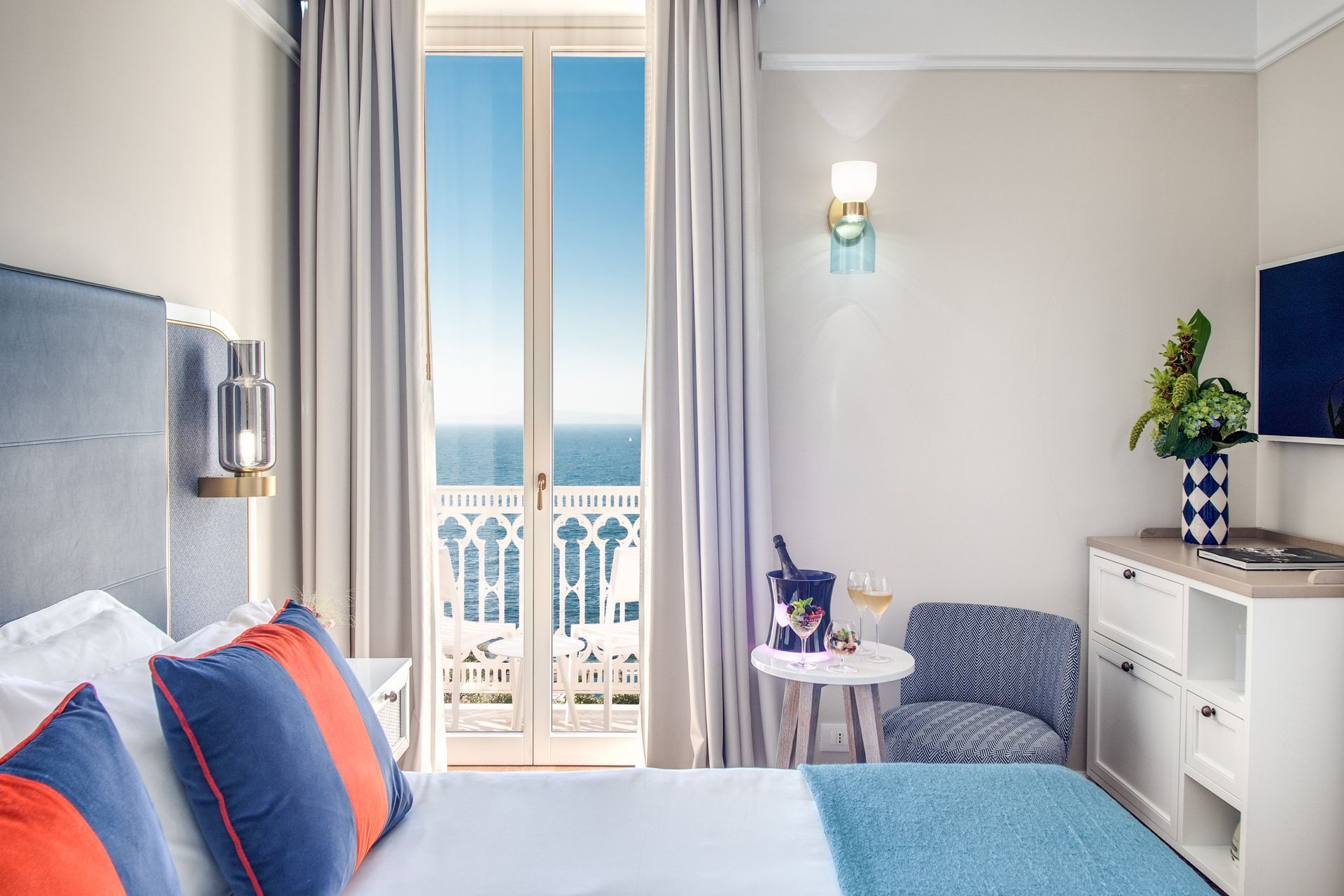 Una camera d'albergo con balcone vista mare, un letto con cuscini blu e arancioni, un tavolino e una poltrona a fantasia.