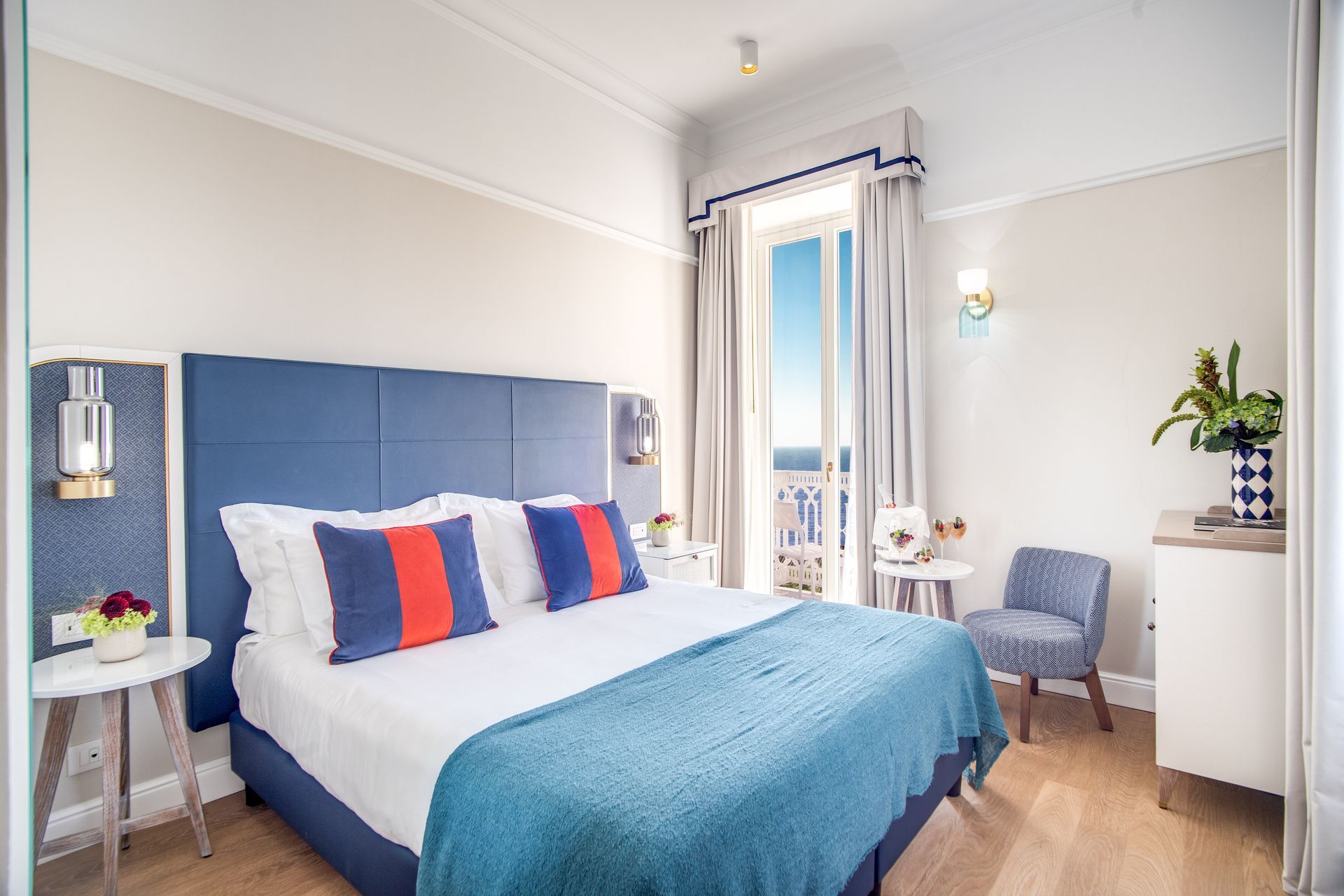 Una moderna camera d'albergo con un letto imbottito blu, cuscini decorativi, un comodino, una sedia a fantasia e vista sul mare.