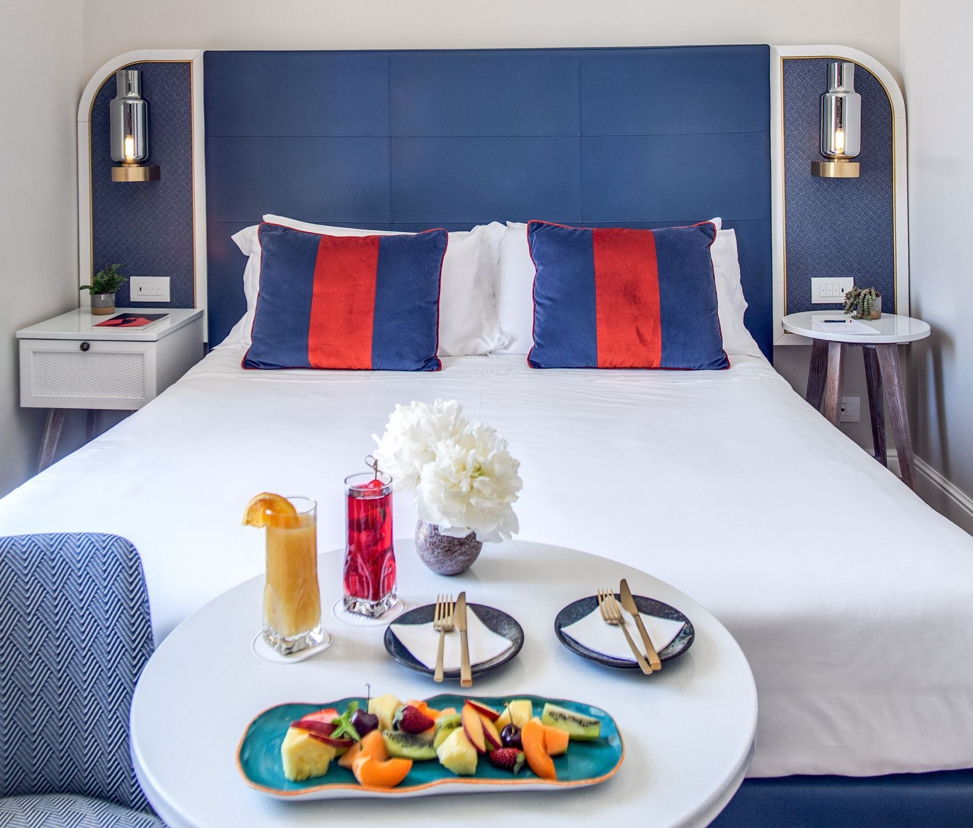 Un letto d'albergo con testiera blu, affiancato da comodini e lampade, con un tavolino rotondo di fronte su cui sono appoggiati frutta e bevande.