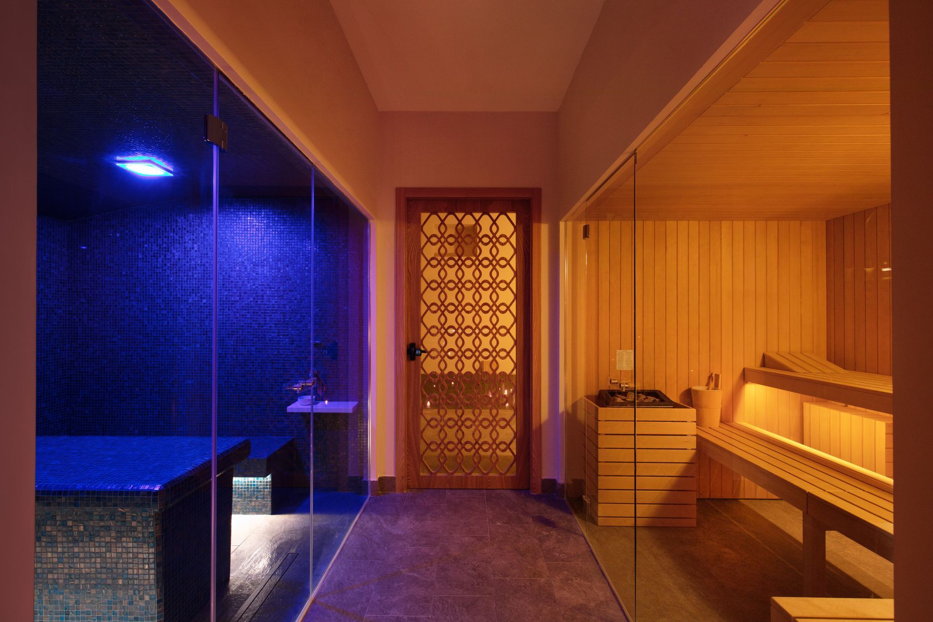 Un corridoio fiancheggiato a sinistra da un bagno turco con piastrelle illuminate di blu e a destra da una sauna in legno con porta decorata.