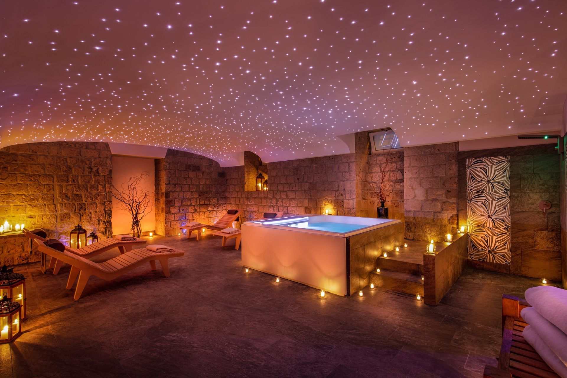 Una tranquilla sala spa con pareti in pietra, vasca idromassaggio illuminata, atmosfera a lume di candela e un effetto luminoso che ricrea un cielo stellato sul soffitto.