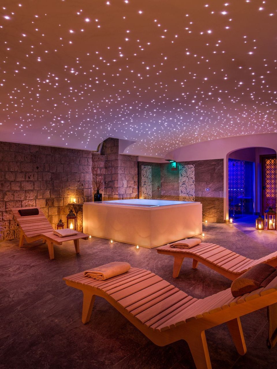 A spa room with stone walls, a square hot tub, and lounge chairs under a ceiling lit with tiny, starry LED lights.