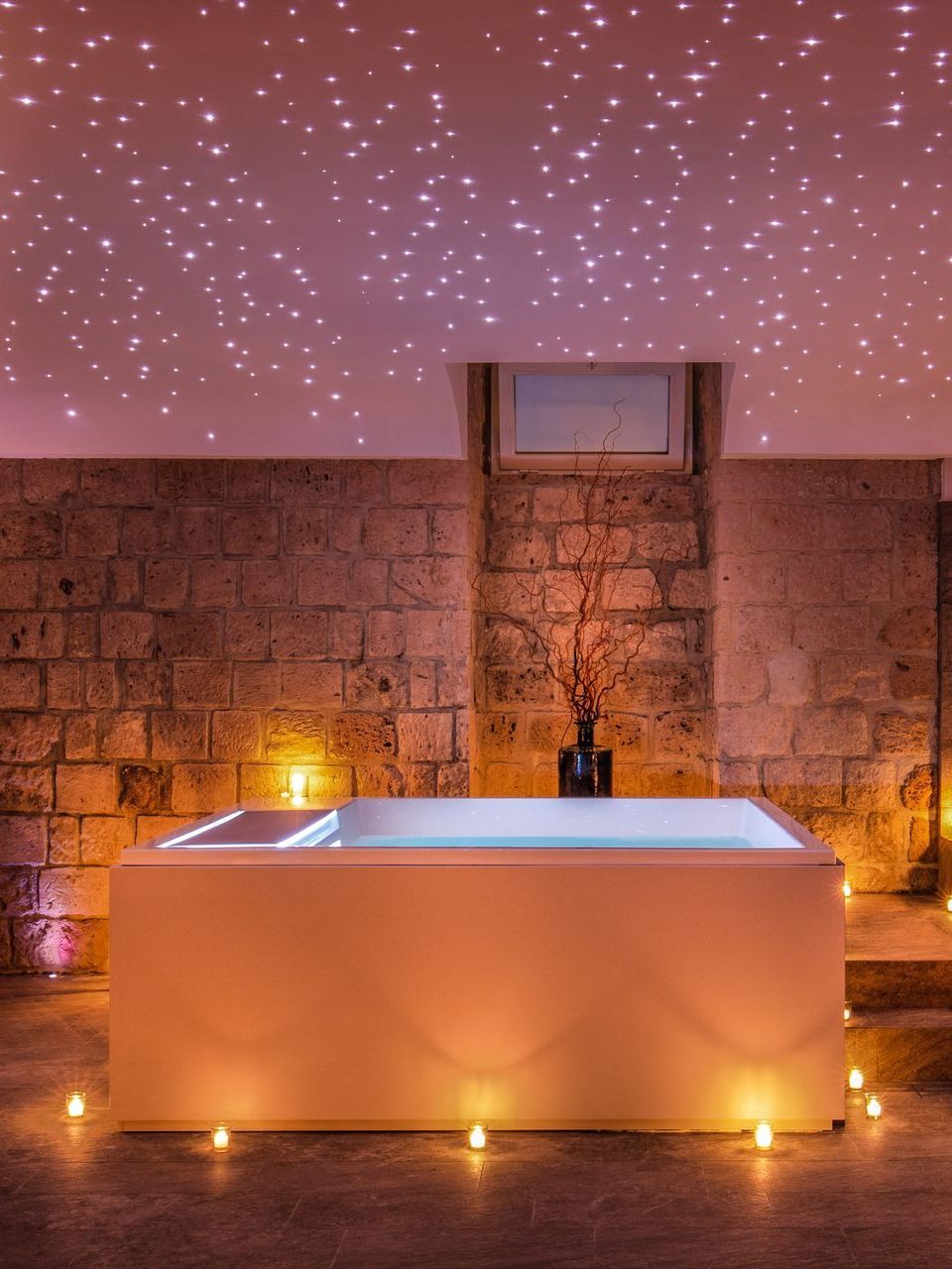 A modern, rectangular bathtub sits in a stone-walled room, illuminated by glowing floor candles and a starry ceiling light.