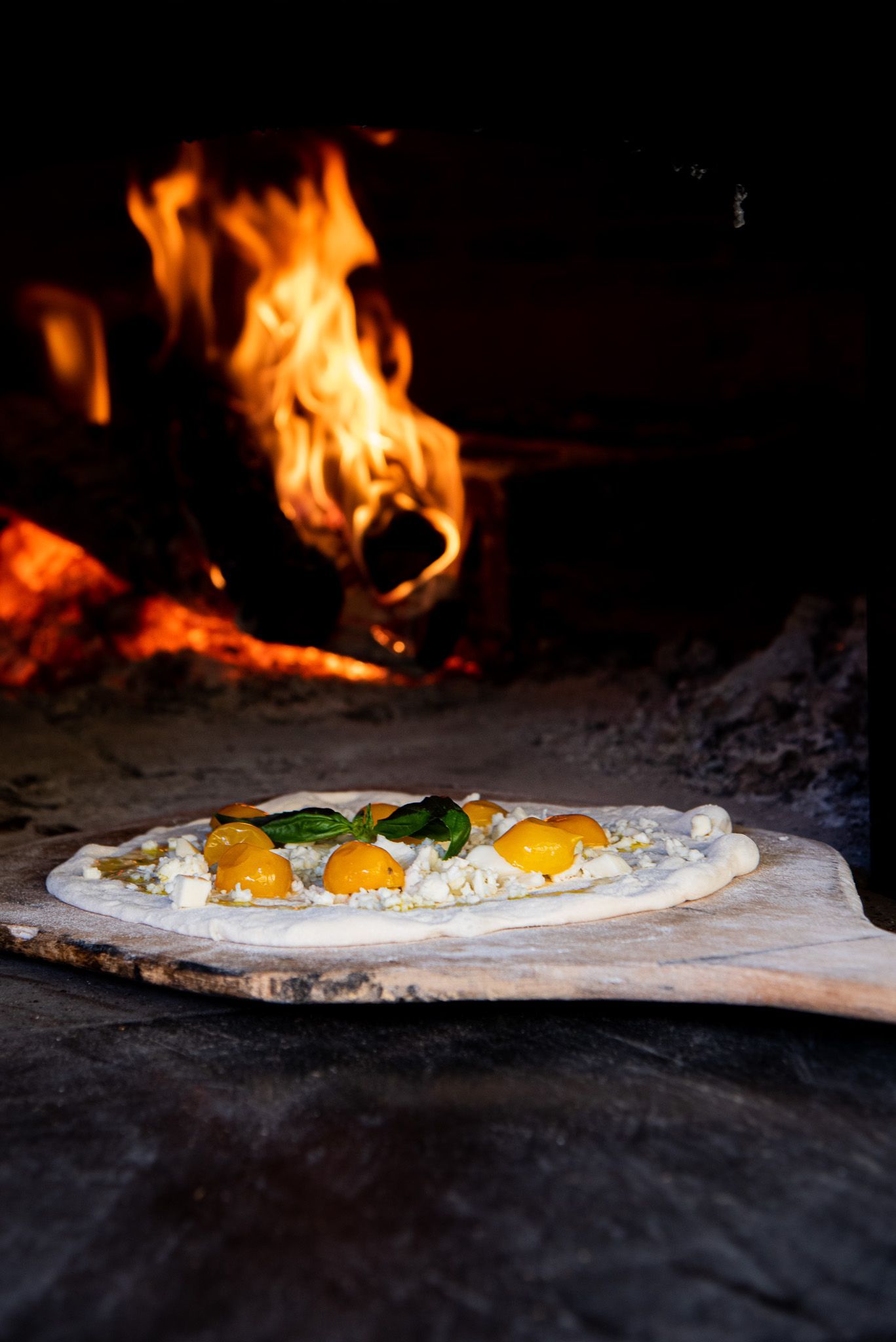 A pizza topped with yellow tomatoes and basil sits on a wooden peel in front of a roaring wood-fired oven.