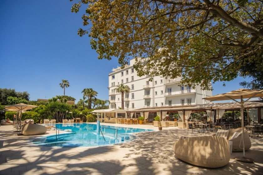 A swimming pool at a bright white resort hotel under a clear blue sky, shaded by large trees and lounge furniture.
