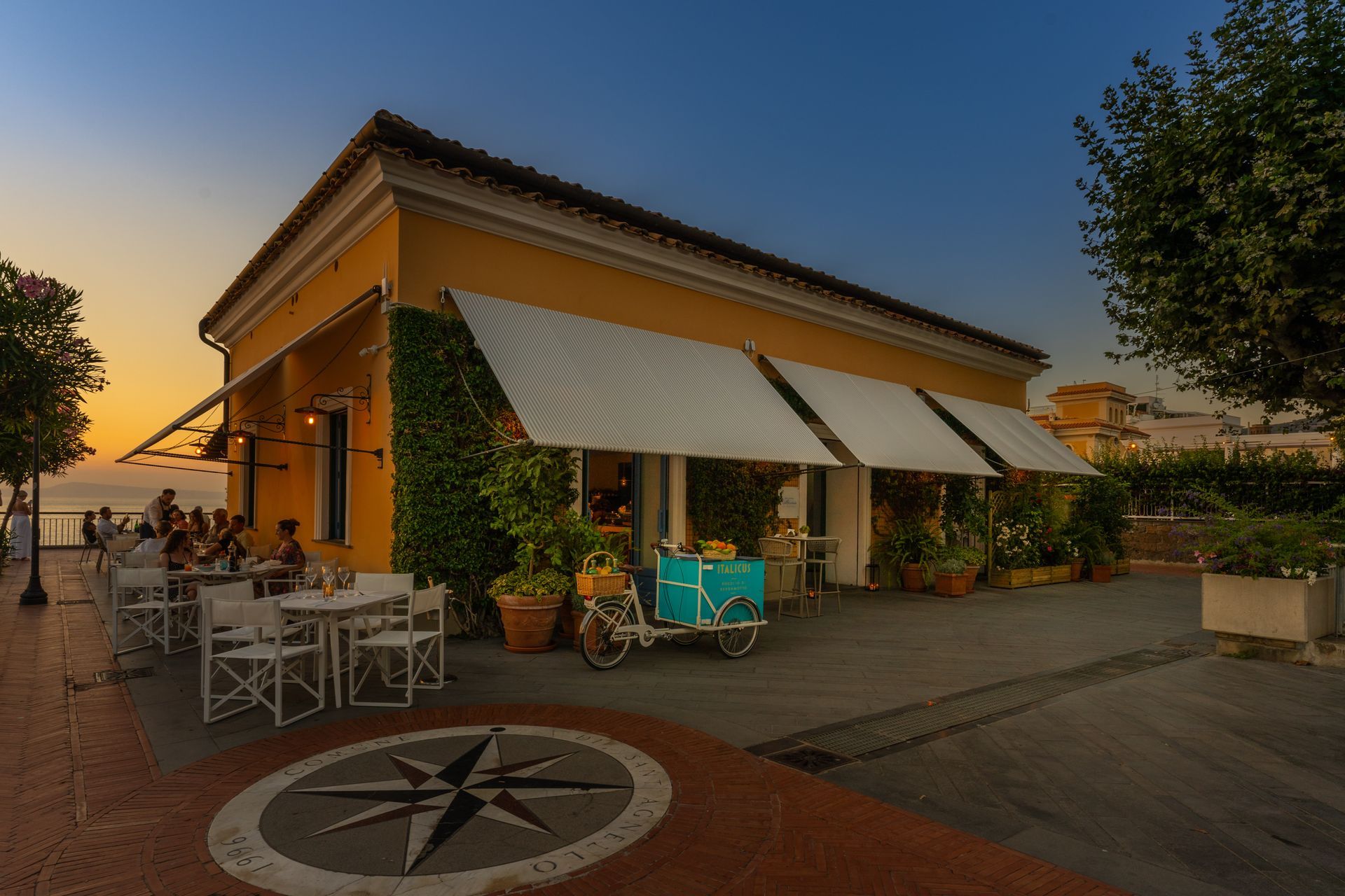 Sunset view of a café patio with awnings, outdoor tables, and a large compass rose in the foreground