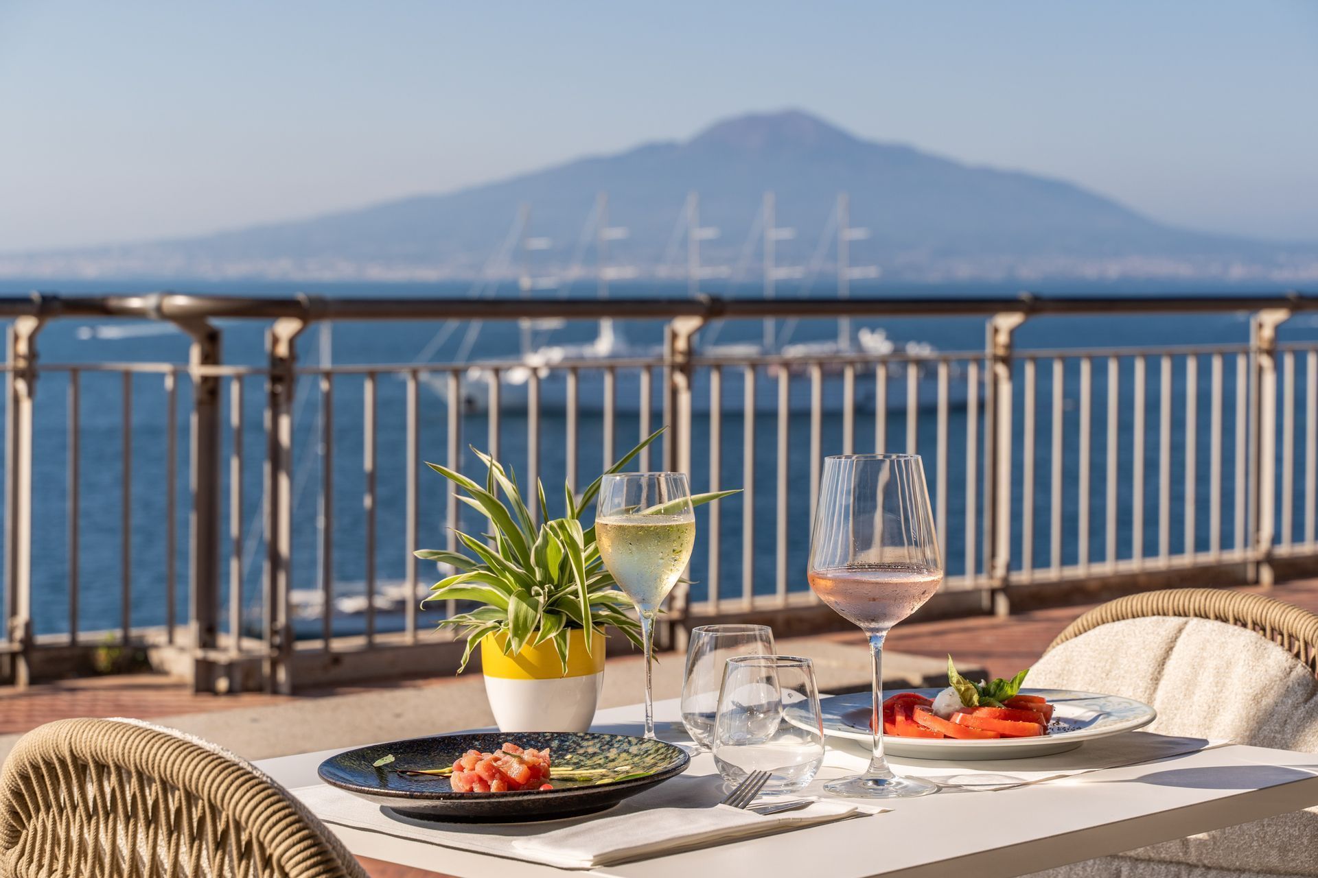 Due bicchieri di vino e piatti di cibo su un tavolo con vista sul mare e sulla montagna, sotto un cielo azzurro e limpido.