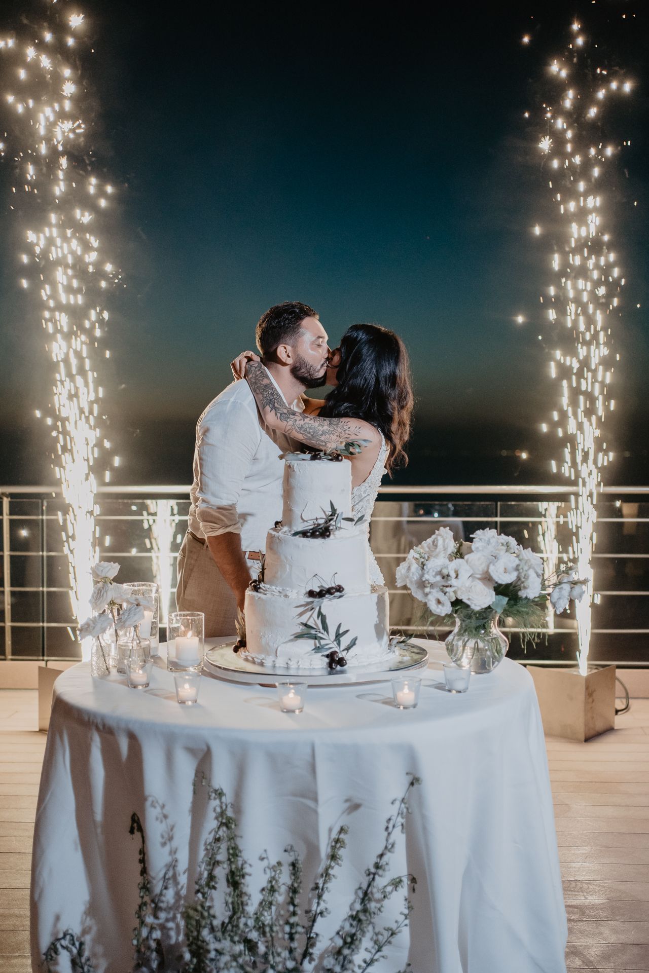 A couple kisses behind a three-tier wedding cake on a terrace at night, framed by two sparkling fountains of light.