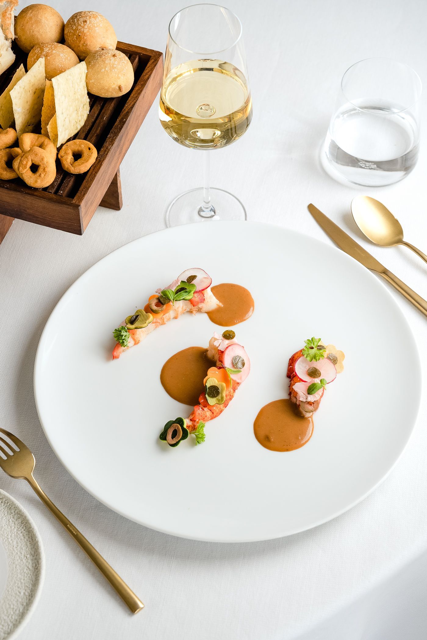 A gourmet dish featuring three small portions of seafood with sauce, served on a white plate with cutlery and wine.