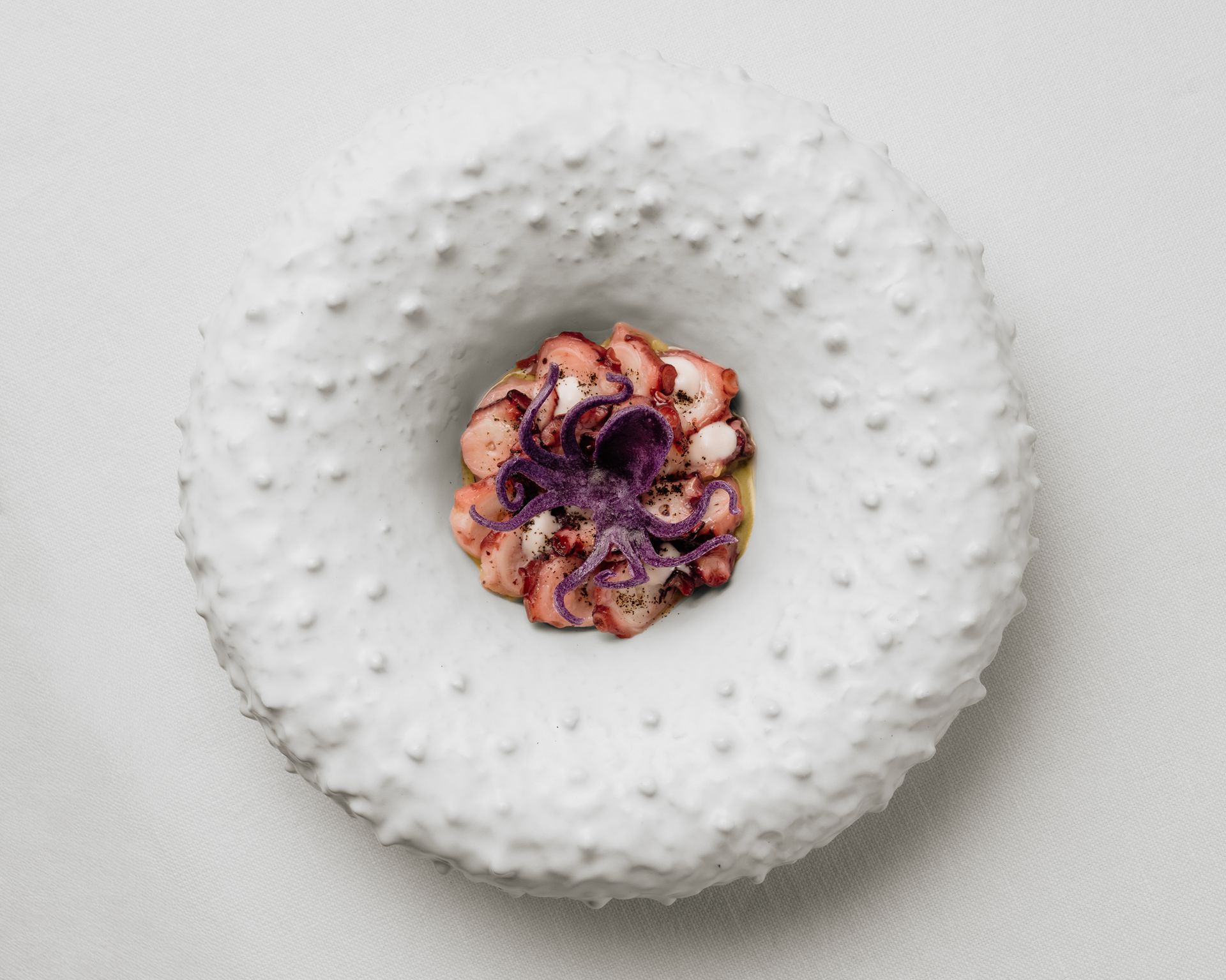 White plate with a ring-shaped white dessert topped with purple berries at the center