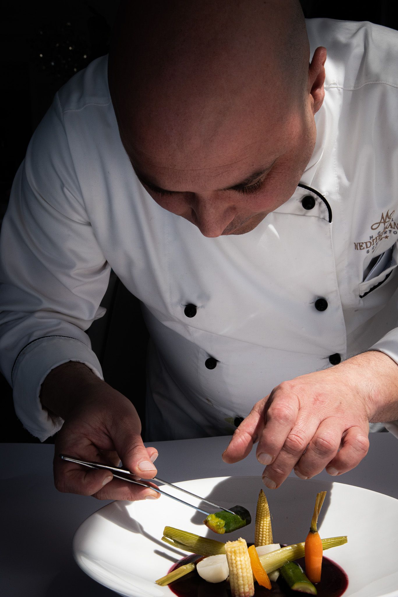 A chef in a white uniform uses tweezers to carefully arrange small, colorful vegetables on a plate.
