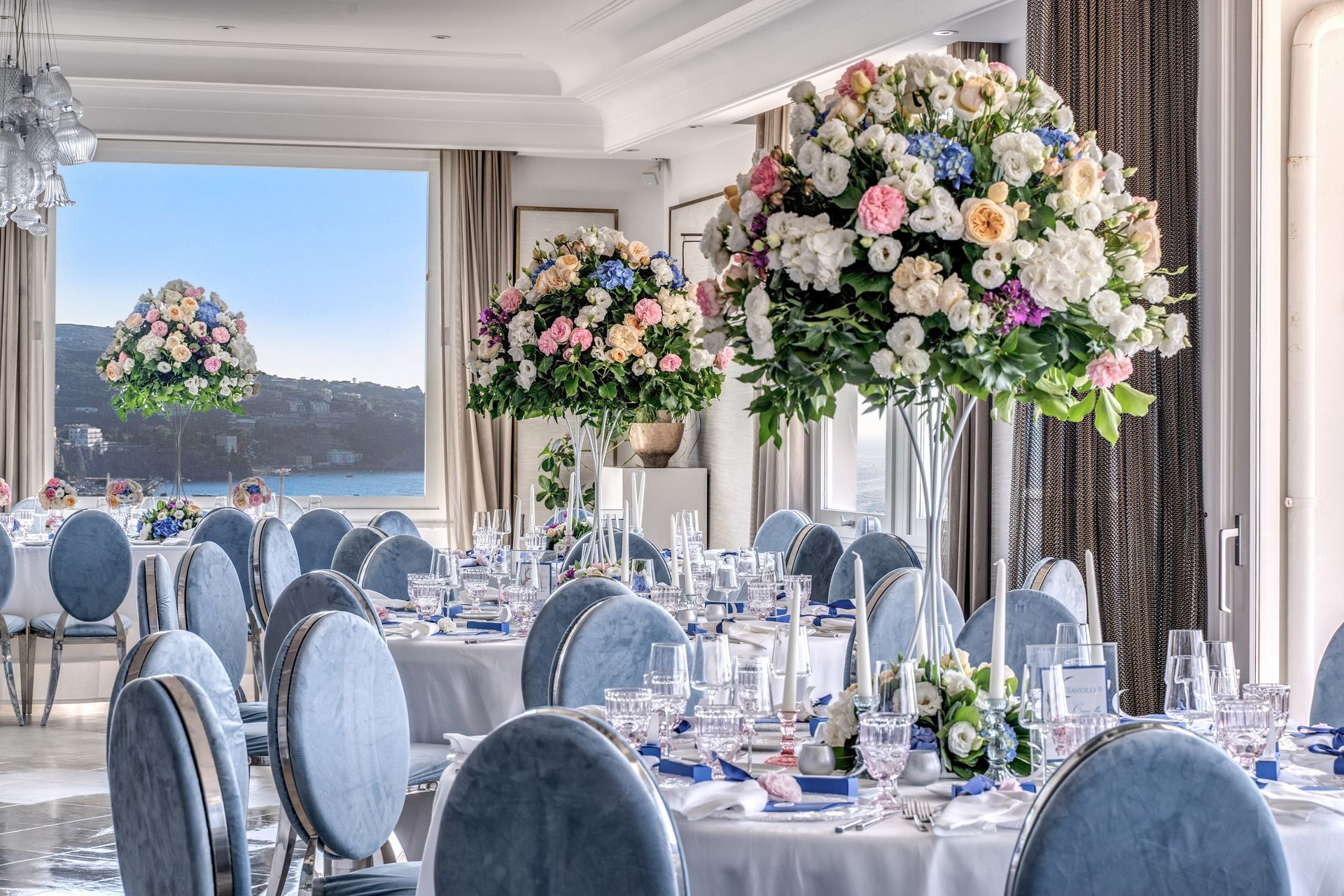 A formal dining room set for a reception with round tables, blue chairs, and large, colorful floral centerpieces.