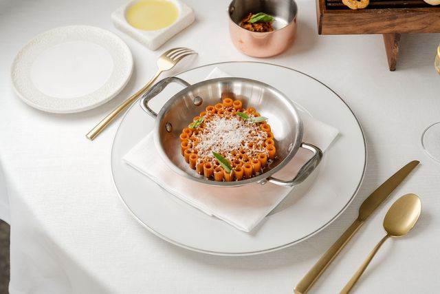 Su un piatto posto su un tavolo bianco, una piccola padella in acciaio inox con pasta al sugo di pomodoro, guarnita con formaggio ed erbe aromatiche.