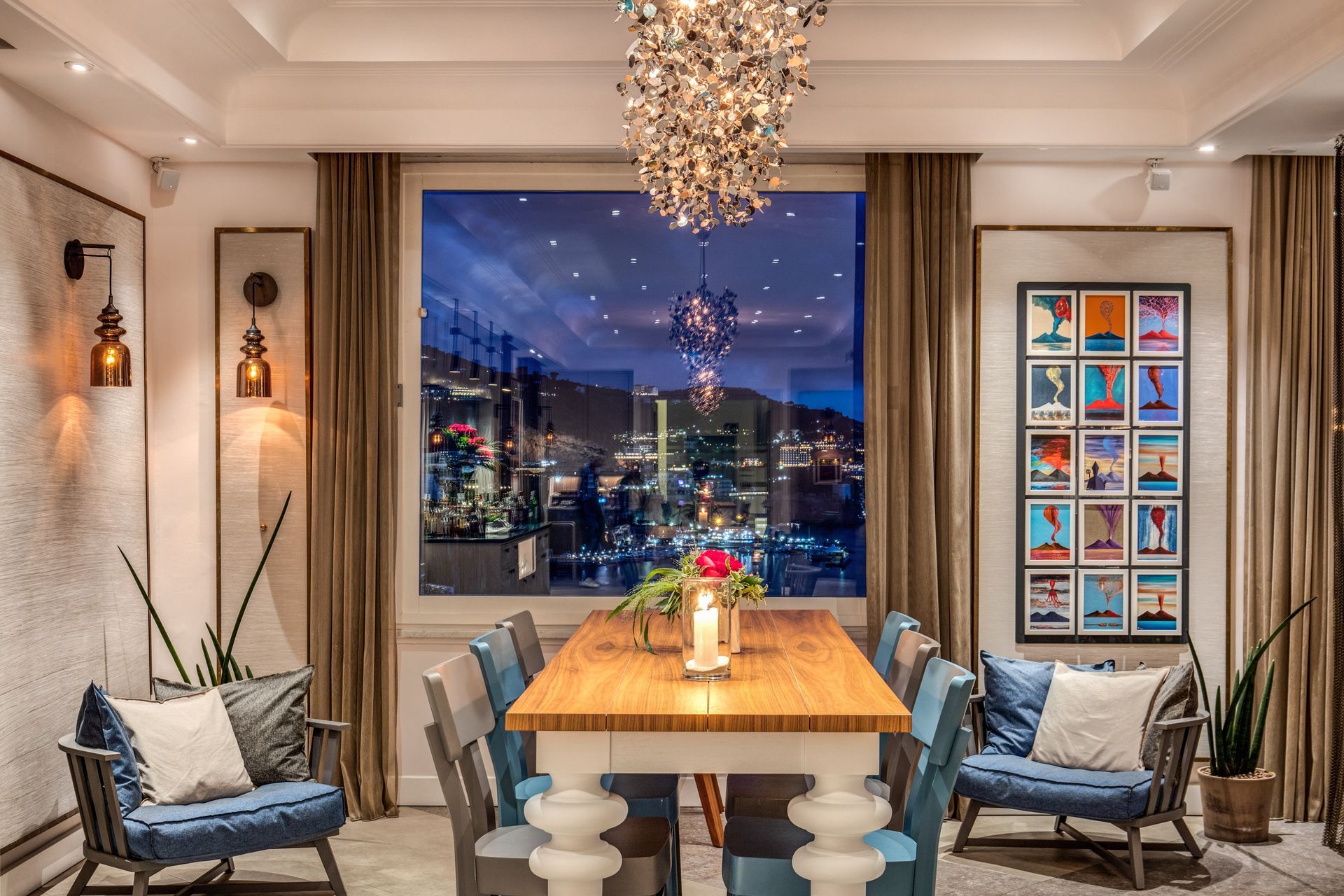 A dining room with a wooden table, blue chairs, a decorative chandelier, and a large window view at night.