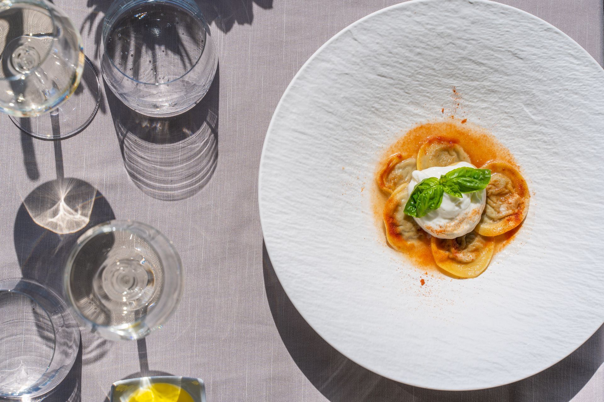 A gourmet pasta dish with cream and basil sits on a textured white plate beside wine glasses on a grey tablecloth.