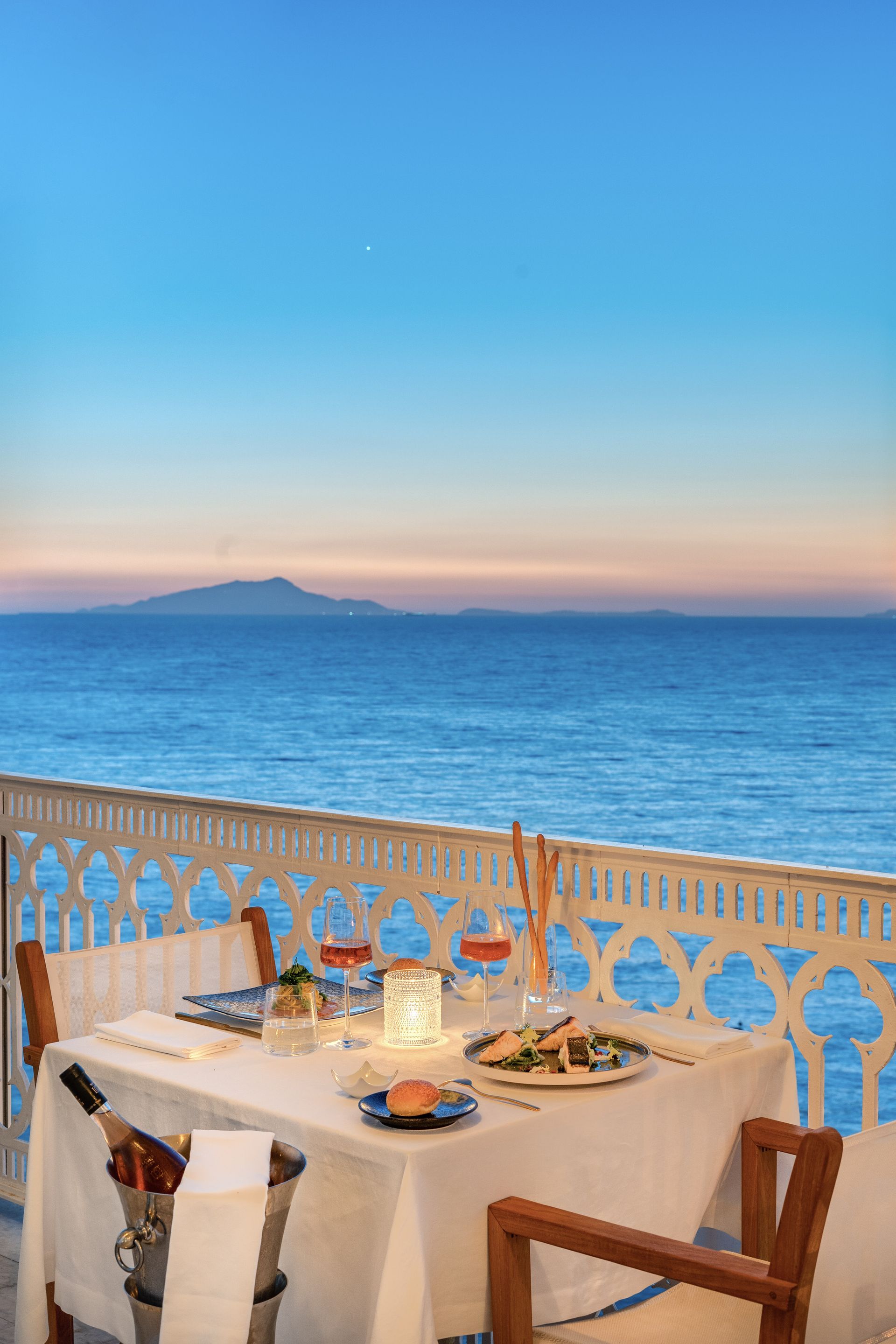 A table set for a romantic dinner on a balcony overlooking the blue ocean at sunset.